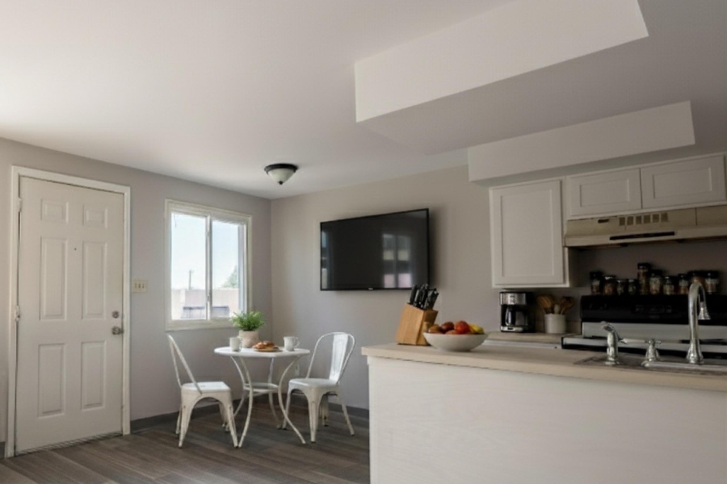 A cozy kitchen and dining area featuring a small round table with two white chairs, a light-colored countertop, and a modern kitchen setup. A window allows natural light, and a flat-screen TV is mounted on the wall, while kitchen appliances and utensils are neatly organized on the counter.