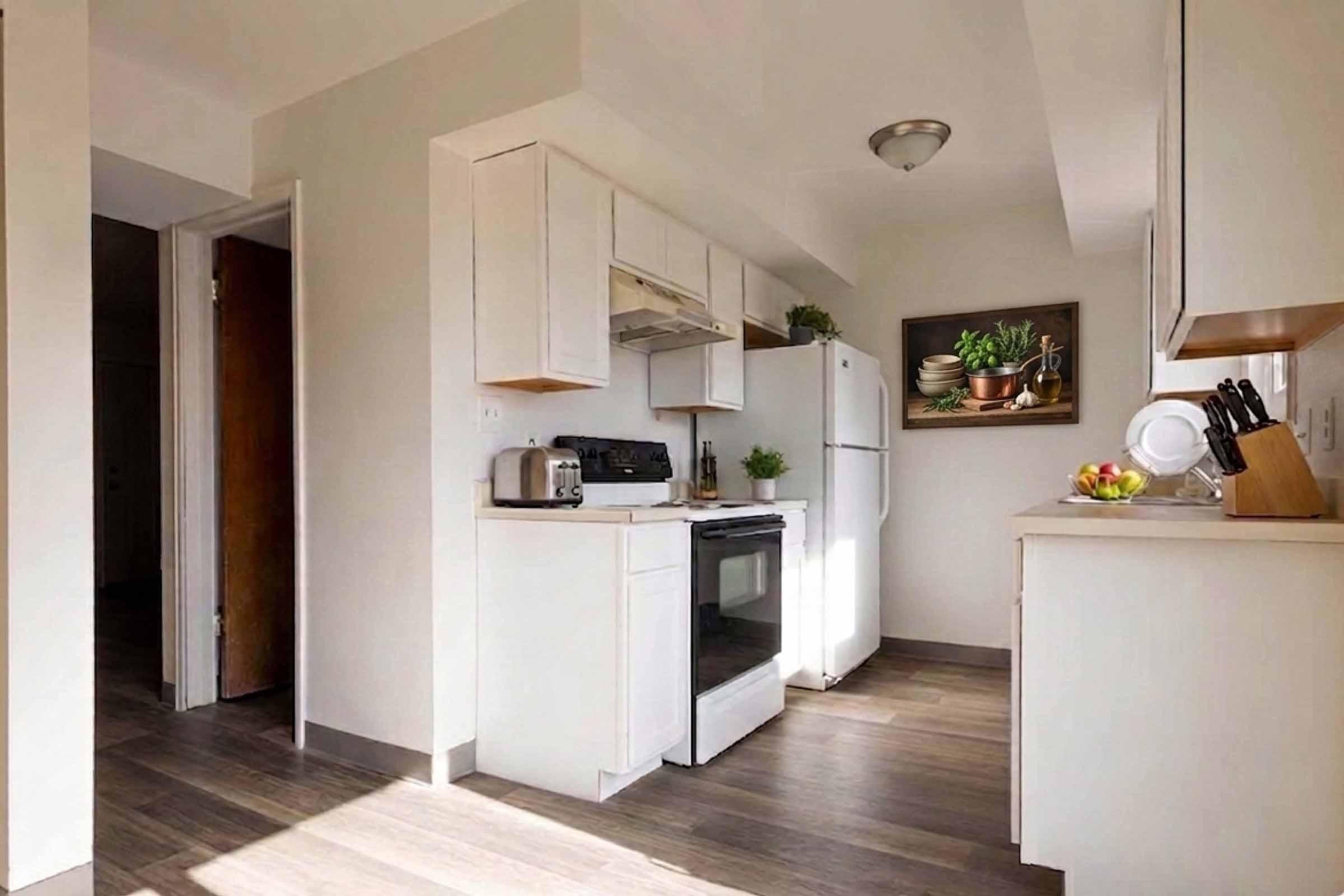 A modern kitchen with white cabinets, a stove, and a refrigerator. The space features light wooden flooring, a small table, and a framed painting of plants on the wall. Countertops hold kitchen items like a kettle and a bowl of fruit, creating a bright and inviting atmosphere.