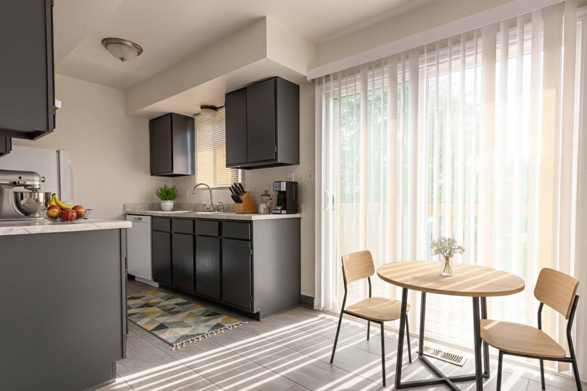 Modern kitchen featuring dark cabinets, a marble countertop, and a small dining area with two wooden chairs and a round table. Natural light streams in through sheer curtains, illuminating a rug on the floor and a bowl of fruit on the countertop. A plant adds a touch of greenery to the space.