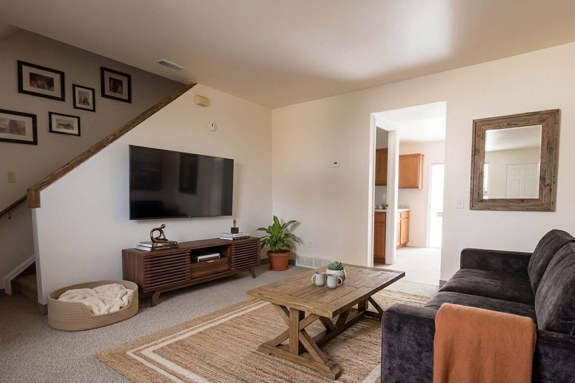 A cozy living room featuring a dark gray sofa, a wooden coffee table, and a large TV mounted on the wall. A potted plant adds a touch of greenery, while a textured rug and warm-toned throw enhance the inviting atmosphere. Framed pictures are displayed on the wall, and a room opens into a kitchen area.