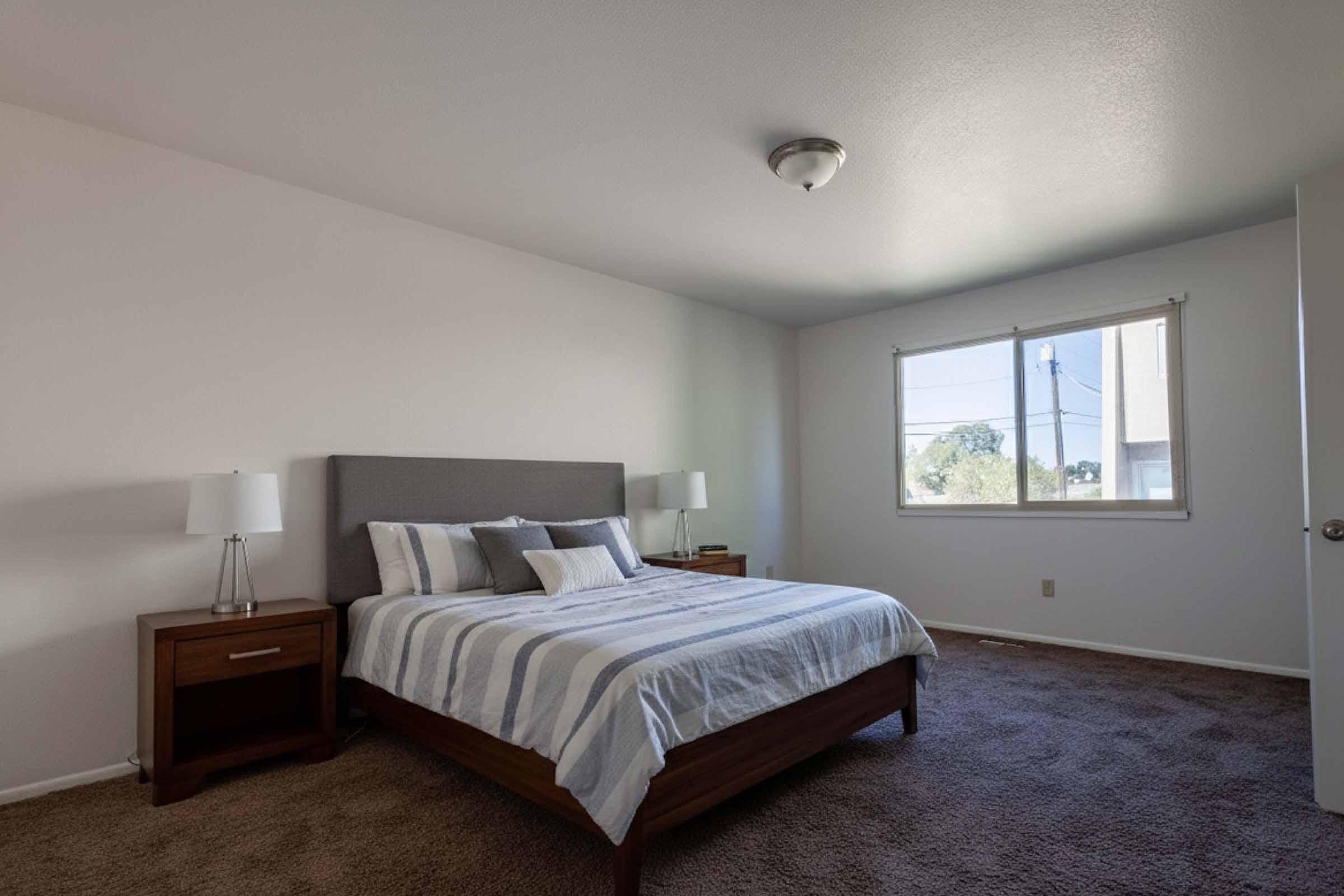 A cozy bedroom featuring a neatly made bed with gray and white striped bedding. The room has light-colored walls, a window with natural light, and wooden nightstands with table lamps on either side of the bed. Soft, plush carpeting adds warmth to the space.