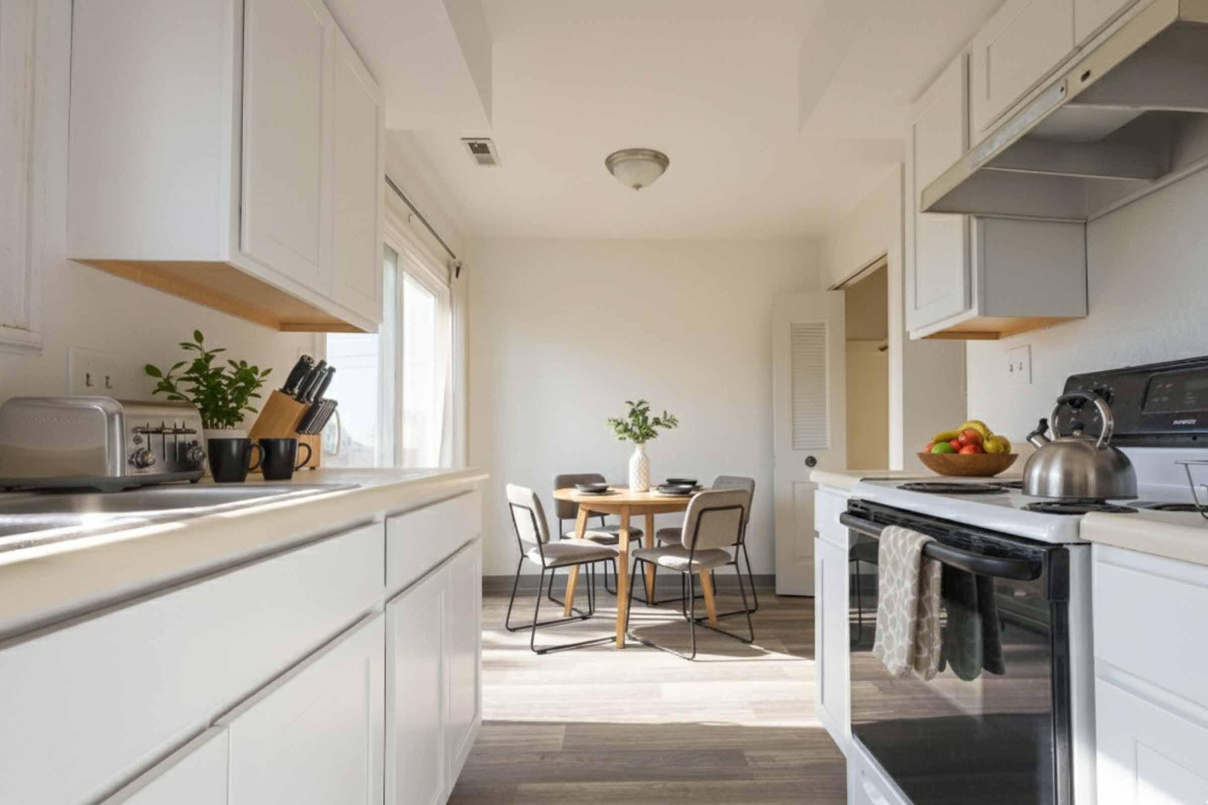 Bright and modern kitchen featuring white cabinets, a sleek black stove, and a light wooden dining table set for two. A bowl of fresh fruit sits on the table, with green plants decoratively placed on the countertop. Large windows allow natural light to fill the space, creating a warm and inviting atmosphere.