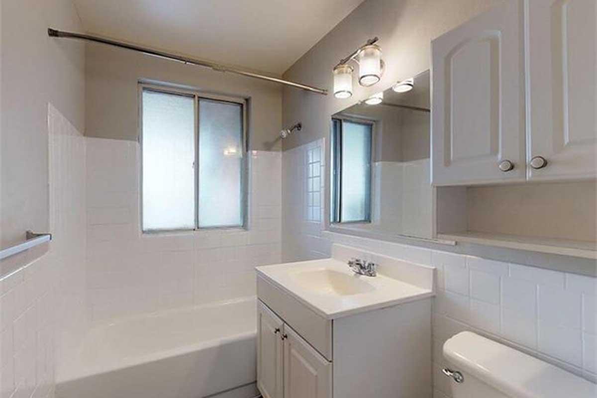 A clean, modern bathroom featuring a white tiled bathtub, a sink with a cabinet underneath, a toilet, and two windows allowing natural light. The walls are painted a light color, and there are three light fixtures above the mirror. The overall appearance is bright and spacious.