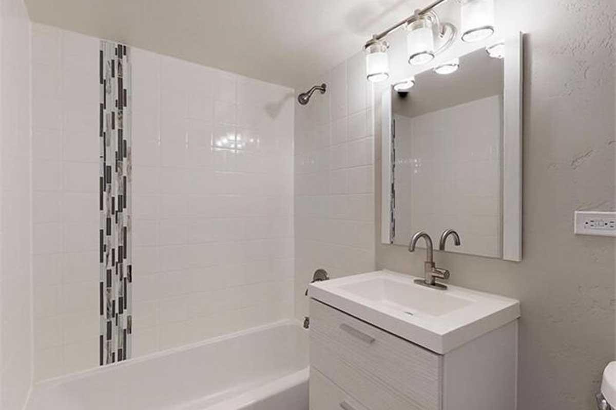 A modern bathroom featuring a white bathtub with a tiled accent wall, a sleek vanity with a basin and faucet, and a large mirror with contemporary lighting above it. The walls are tiled with white ceramic tiles, creating a clean and bright atmosphere.