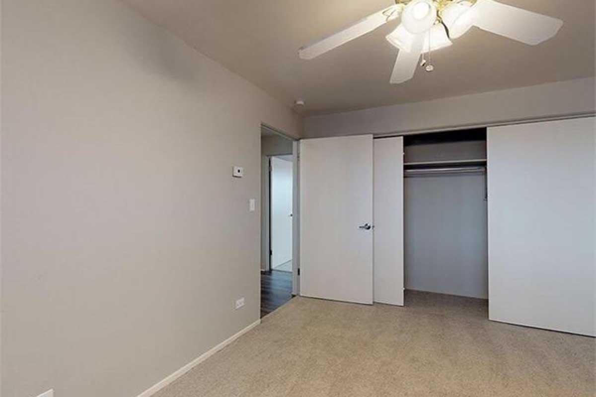 A spacious room with light gray walls and a ceiling fan. There is a closet with sliding doors on one side and a doorway leading to another area. The floor is carpeted in a neutral tone, creating a calm and inviting atmosphere.