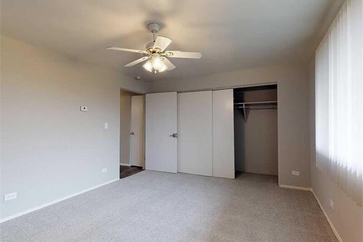 A spacious, empty bedroom featuring light-colored walls and a ceiling fan. The floor is carpeted, and there are two sliding closet doors on one side. A doorway leads to another room, and large windows allow natural light to brighten the space.