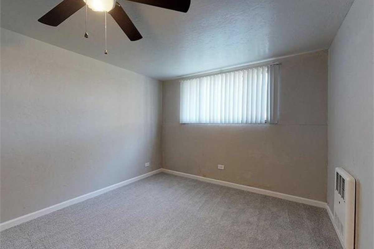 Empty room with light gray carpet, a ceiling fan with light, and a large window covered with white vertical blinds. The walls are painted a light neutral color, creating a bright and airy atmosphere. There is a heating unit on one wall, and the space is free of furniture.