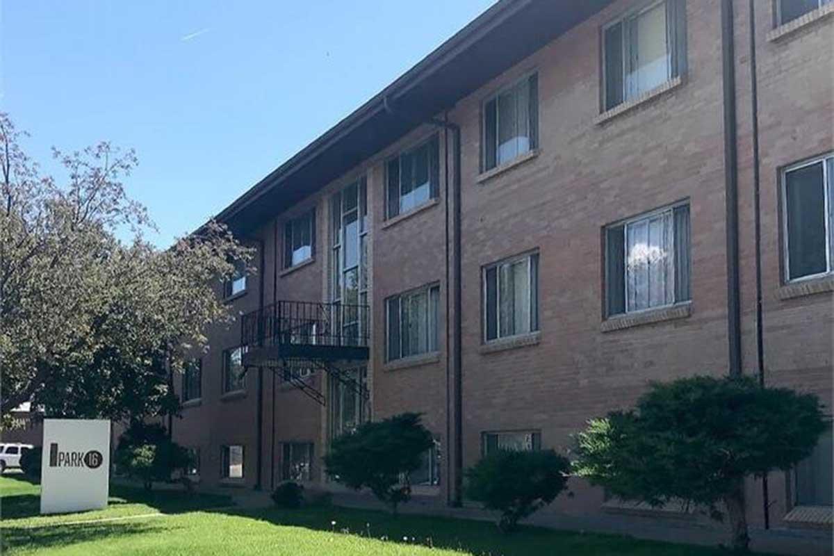 A brick apartment building with multiple windows, featuring a metal spiral staircase on one side. In front, there is a sign that reads "Apartments" surrounded by a well-maintained grassy area and small shrubs. The sky is clear and blue, adding to the bright atmosphere of the scene.