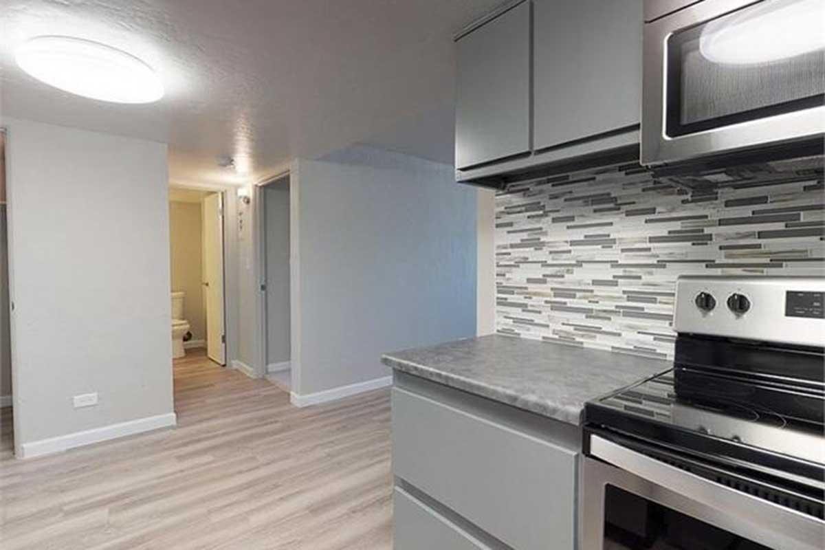 A modern kitchen with gray cabinetry and a stainless steel stove. A stylish backsplash features a mix of gray and white tiles. The adjacent room is brightly lit, showcasing light-colored flooring and a doorway leading to a bathroom. Overall, the space appears clean and contemporary.