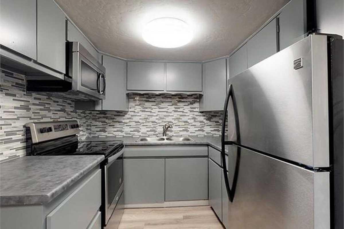 Modern kitchen featuring gray cabinetry, stainless steel appliances including a refrigerator and stove, and a sleek backsplash with a mix of gray tiles. The space is well-lit with a circular ceiling light, complemented by a countertop and wooden flooring.