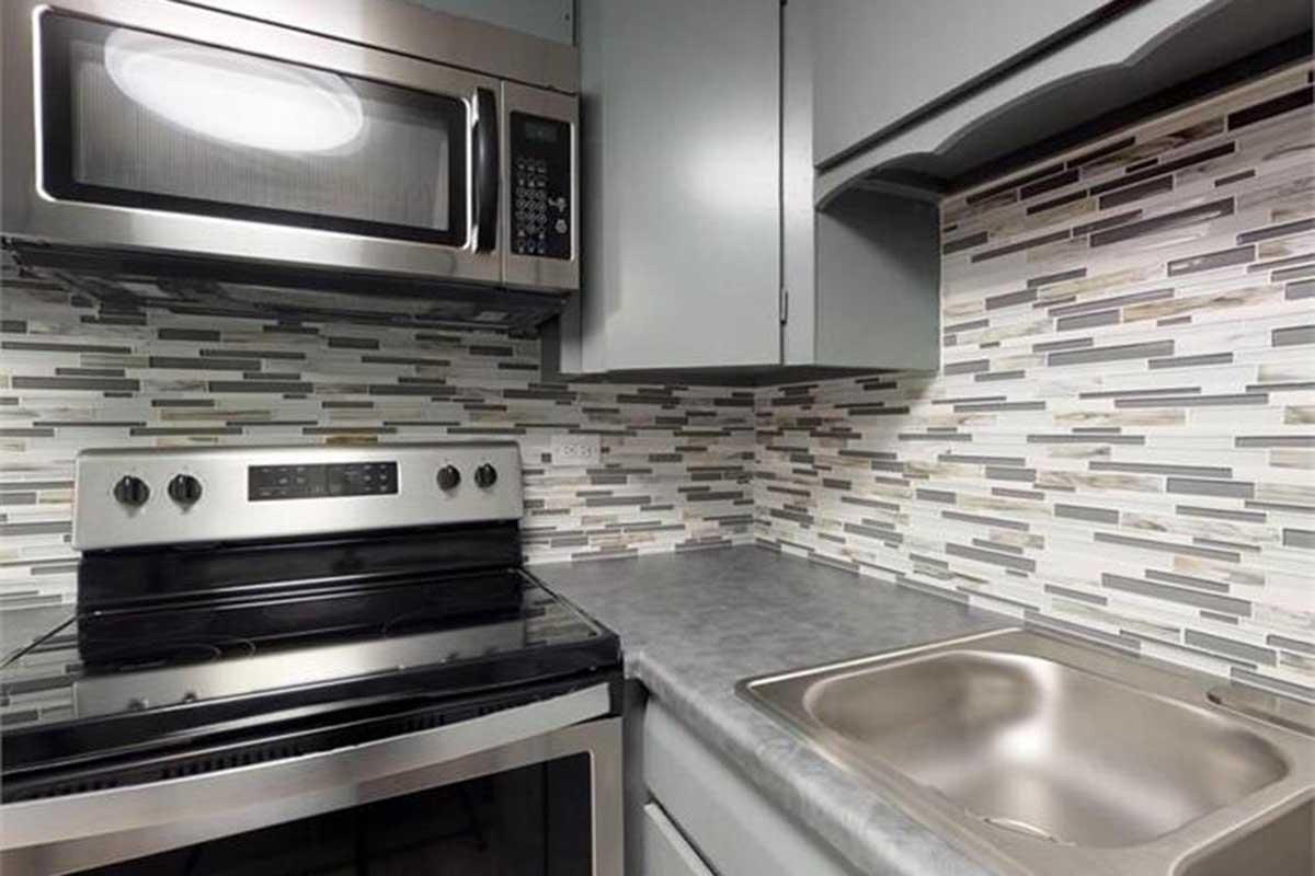 Modern kitchen featuring stainless steel appliances, including a microwave and stove. The backsplash consists of a stylish mosaic of gray and silver tiles, complemented by sleek gray cabinets and a stainless steel sink. The countertop is smooth and contemporary, enhancing the overall aesthetic.