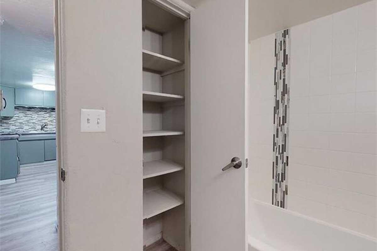 A partially open door reveals a built-in closet with shelves, adjacent to a bathroom featuring a tub and a tiled wall. The setting has neutral colors and modern finishes, with a glimpse of a kitchen area in the background. The floor is light-colored wood.