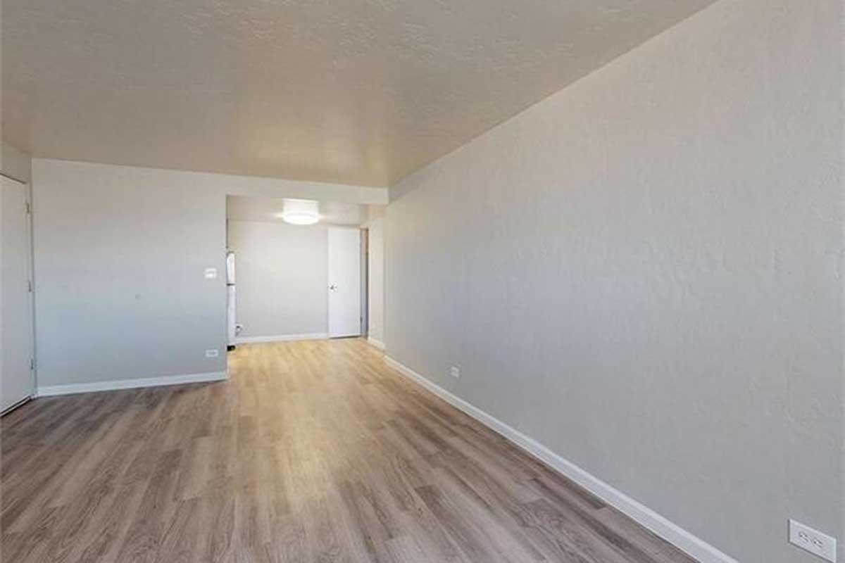 A spacious, empty room with light-colored walls and wooden flooring. A doorway leads to another room, and there is a ceiling light fixture visible. The area is bright and uncluttered, suggesting a modern and minimalist design.