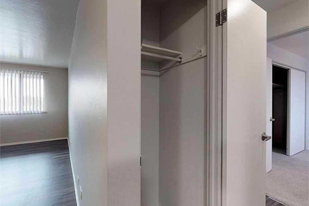 Interior view of a room featuring an open closet with a single rod for hanging clothes. The walls are painted in neutral tones, and there is a window with vertical blinds allowing natural light to enter. The flooring appears to be dark and modern.