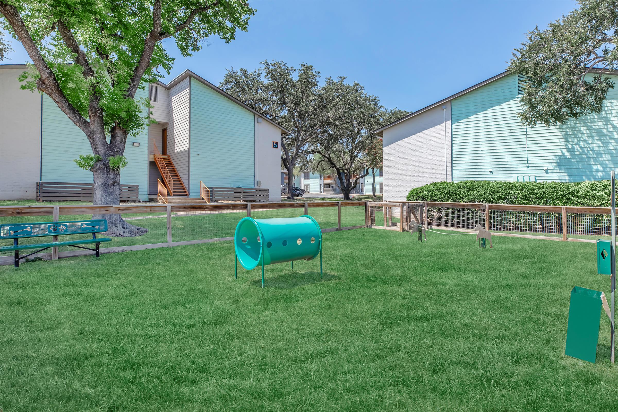 A sunny outdoor area featuring a grassy space with a teal agility tunnel, a fence, and benches for seating. Two light-colored buildings are visible in the background, surrounded by trees and shrubs. The scene conveys a welcoming atmosphere, ideal for recreational activities or relaxation.