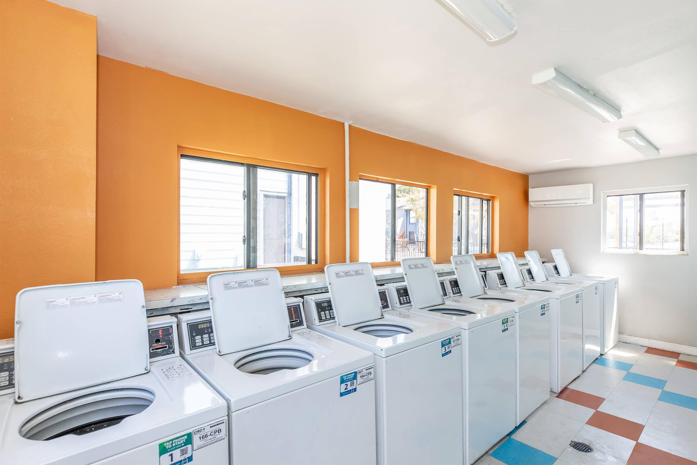 A clean and bright laundromat featuring multiple washing machines lined up against an orange and white wall. Large windows allow natural light to fill the space, enhancing the cheerful atmosphere. The floor has a colorful tiled pattern, creating a welcoming environment for laundry tasks.