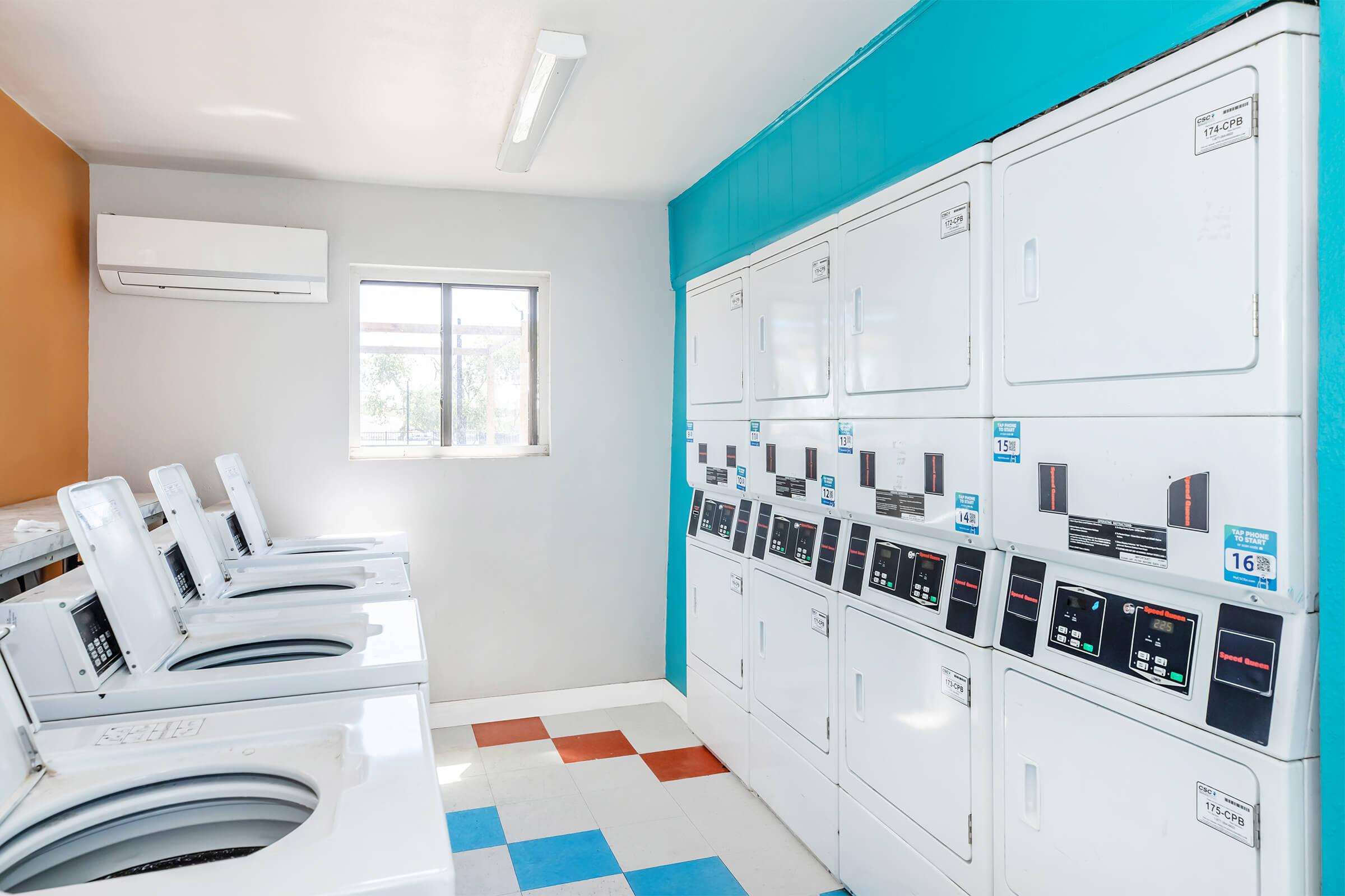 A laundry room featuring several white washing machines and dryers lined up against the wall. The walls are painted in a combination of light blue, orange, and gray. There is a window providing natural light, and the floor is tiled in a checkerboard pattern of blue, white, and red.
