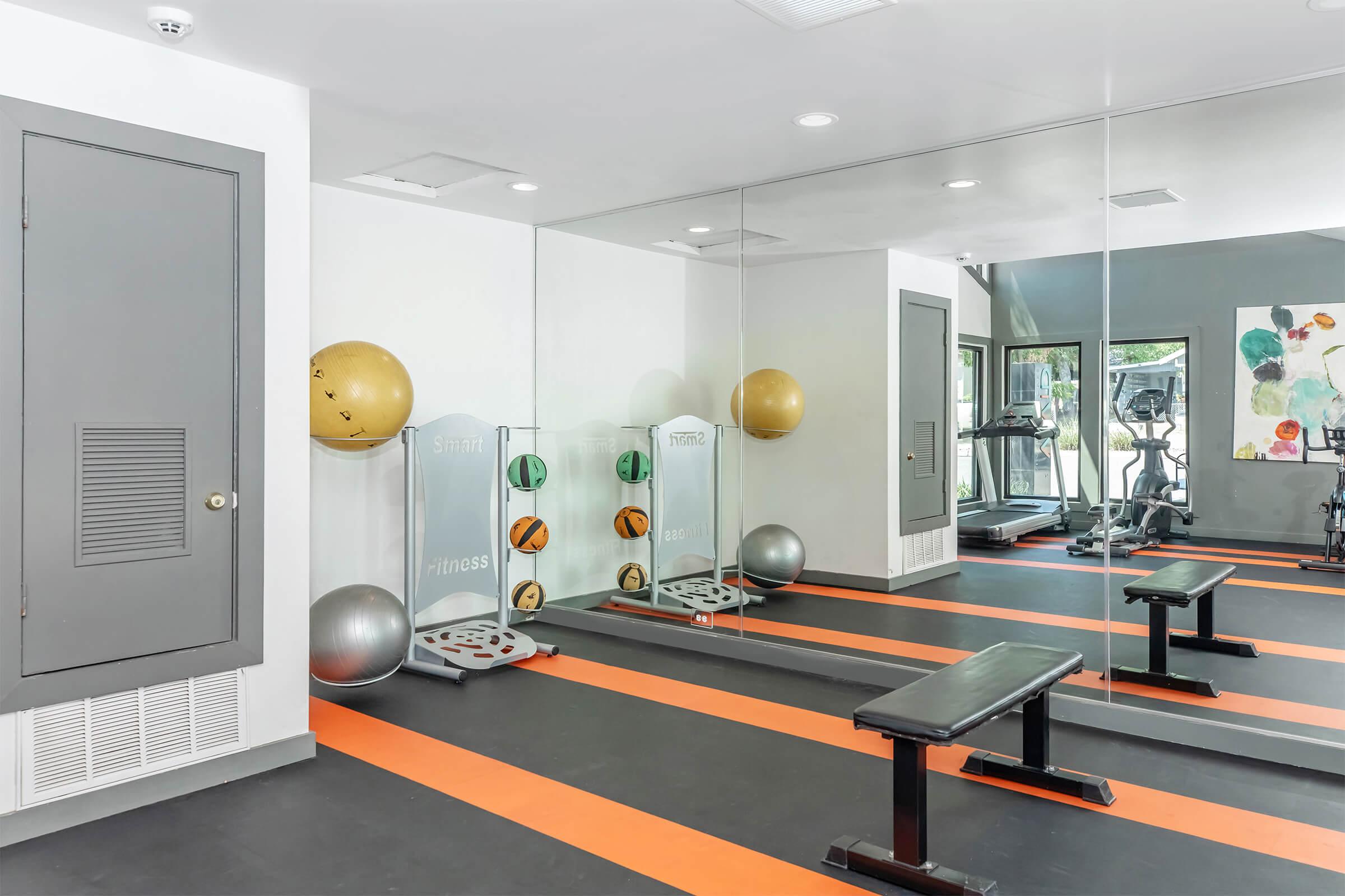 A modern gym interior featuring a mirrored wall, exercise benches, colorful stability balls, and fitness equipment. The floor has orange lines, and there are large windows letting in natural light.