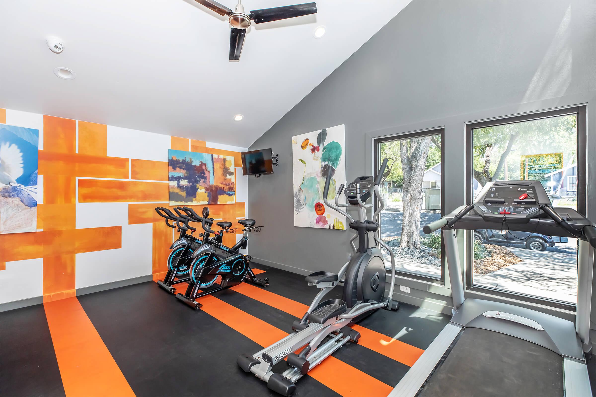 A modern fitness room featuring exercise equipment including stationary bikes, a treadmill, and an elliptical machine. The walls are adorned with colorful artwork, and large windows provide natural light, creating an inviting workout space.