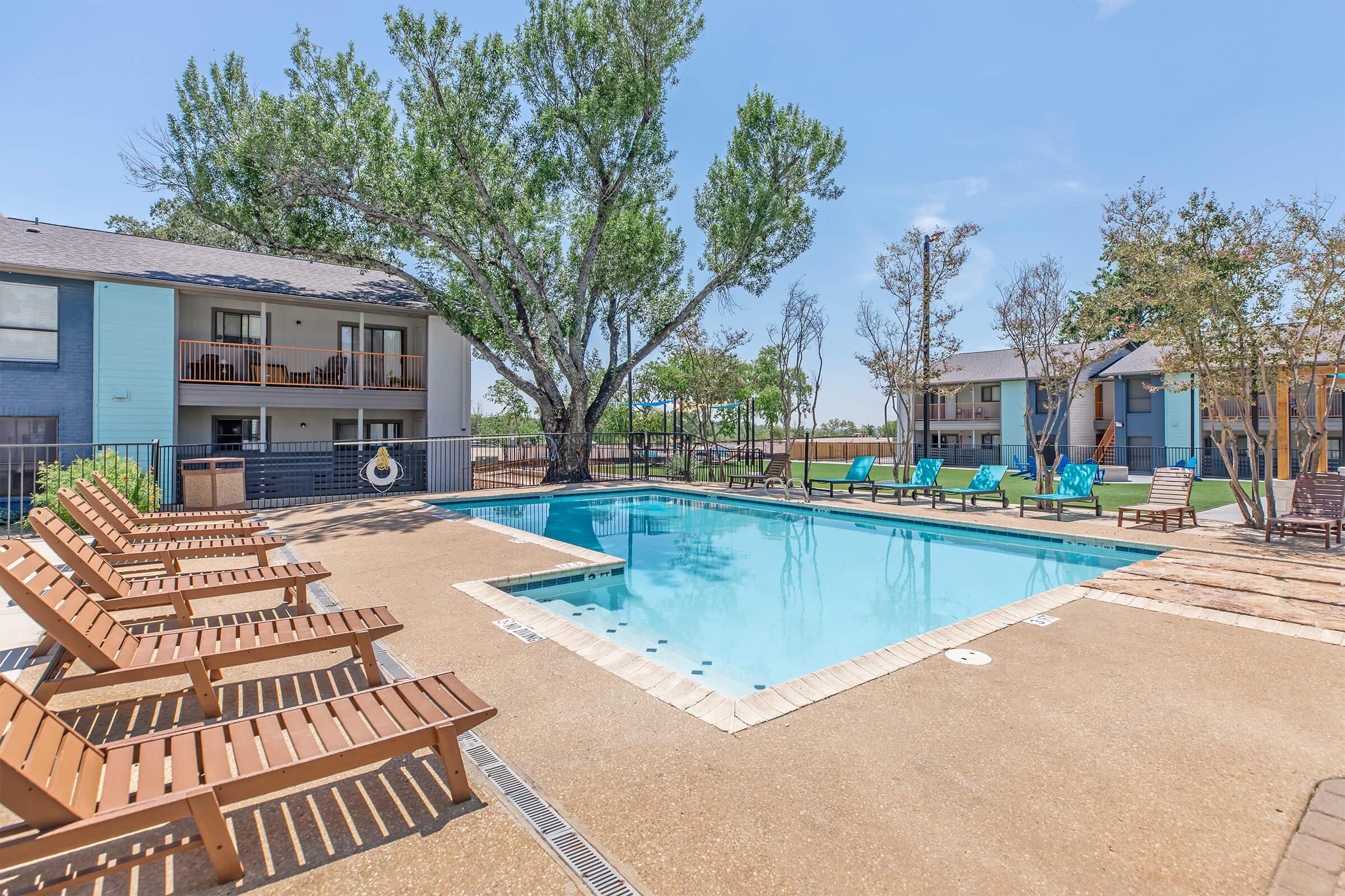 A serene pool area featuring a clear blue swimming pool surrounded by lounge chairs. In the background, there are trees and two multi-story apartment buildings with balconies. The sky is clear and sunny, enhancing the inviting atmosphere of the space.