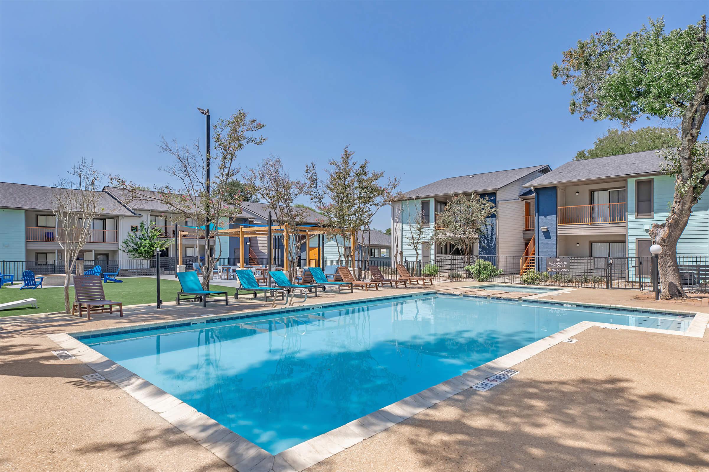 A bright outdoor swimming pool surrounded by lounge chairs and shaded areas. In the background, there are modern apartment buildings with balconies, trees, and a clear blue sky. The setting conveys a sunny, inviting atmosphere, ideal for relaxation.
