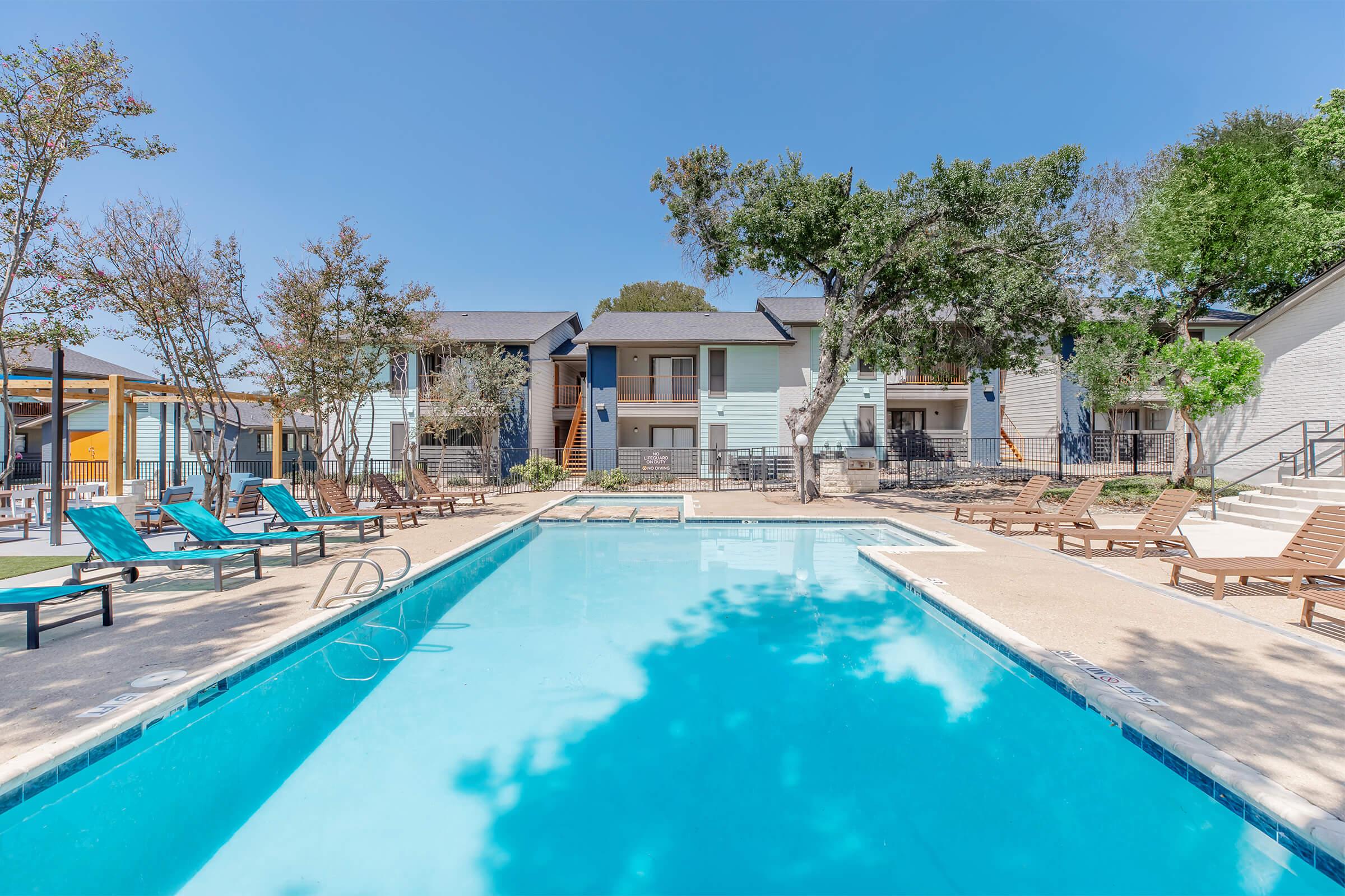 A clear blue swimming pool surrounded by lounge chairs and umbrellas, with a backdrop of modern apartment buildings and trees under a bright blue sky. The scene conveys a relaxing outdoor setting perfect for leisure and socializing.
