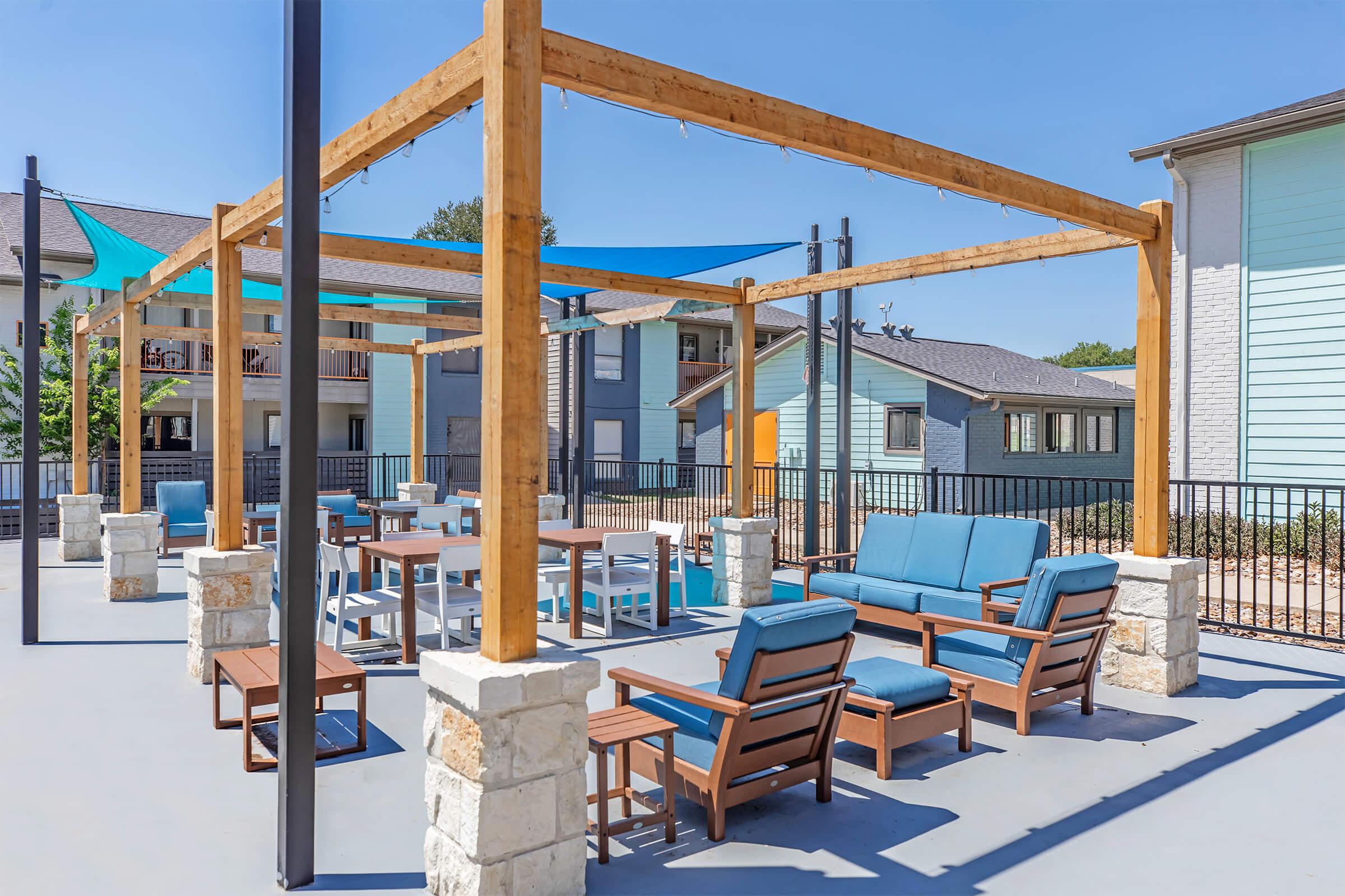 Outdoor lounge area featuring wooden pergolas, comfortable seating, and tables. The space is surrounded by buildings and is designed for relaxation and socializing. Bright blue skies and shaded areas enhance the inviting atmosphere.