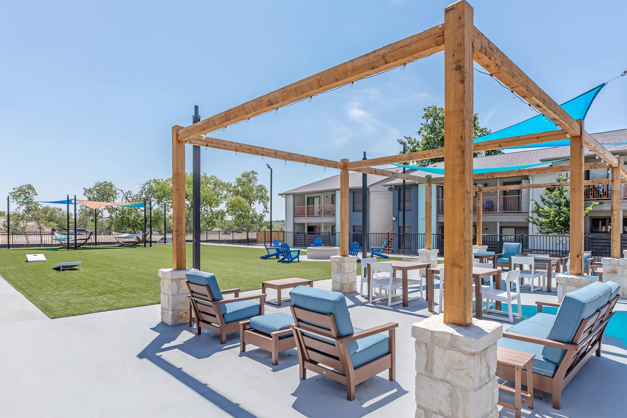 A modern outdoor lounge area featuring wooden pergolas, comfortable seating, and tables. A grassy space with recreational activities, including a volleyball court in the background, and trees providing shade. The atmosphere is bright and inviting, ideal for relaxation and social gatherings.