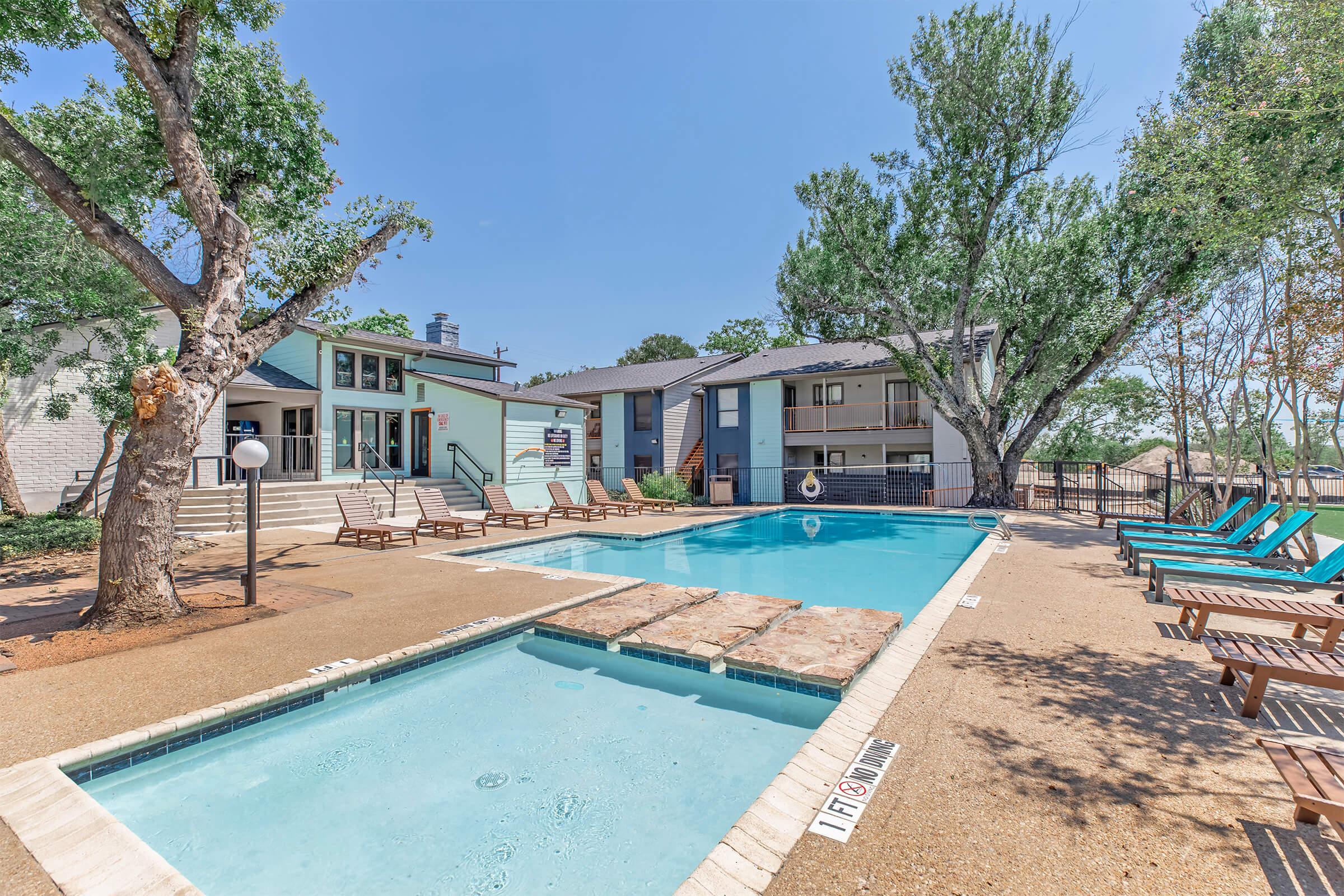 Swimming pool area featuring a large pool with a shallow section, surrounded by lounge chairs. In the background, there are residential buildings and trees providing shade. The scene is set on a clear blue sky day, creating a relaxing outdoor atmosphere.