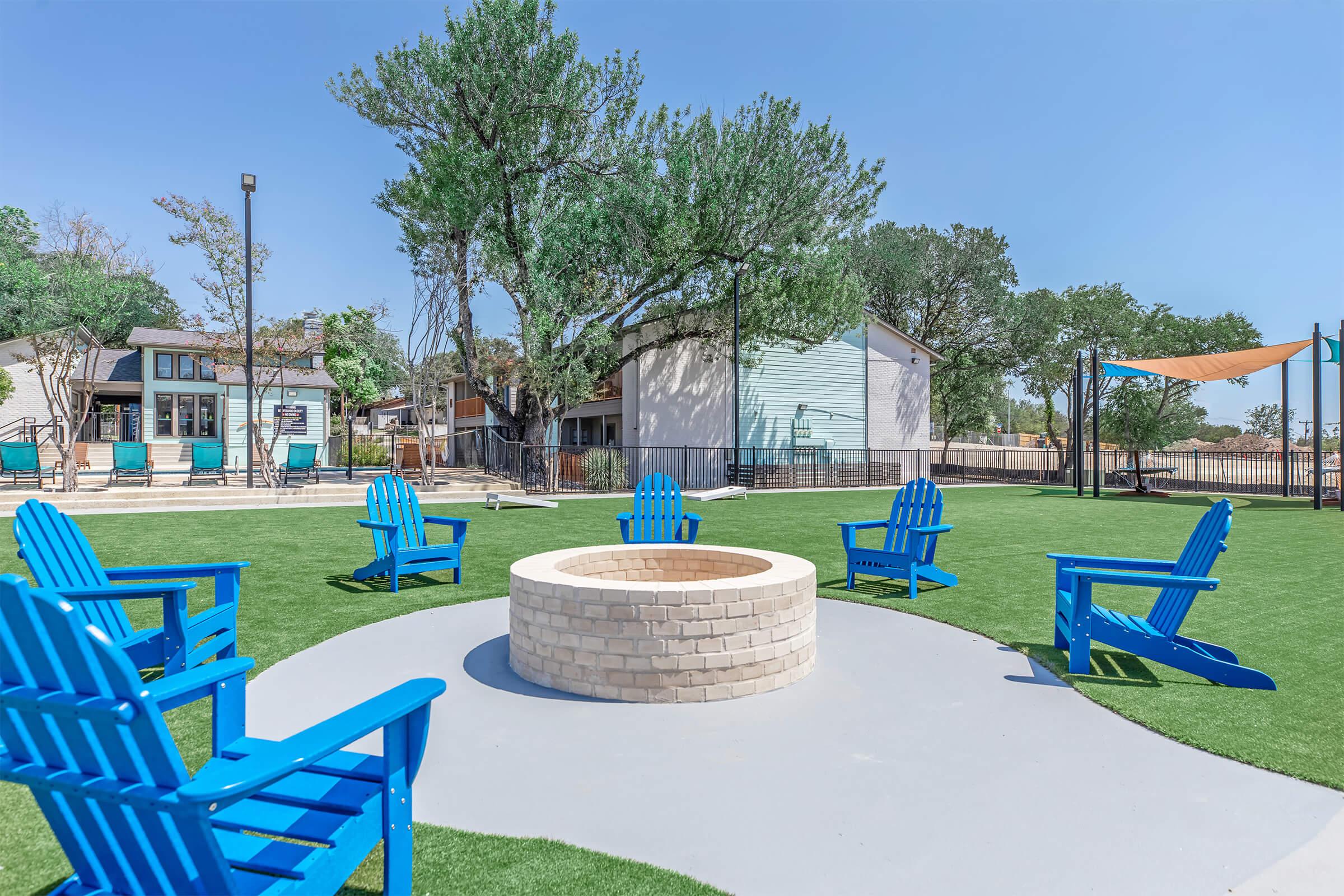 A circular stone fire pit surrounded by bright blue Adirondack chairs on a grassy area. In the background, there are residential buildings and trees under a clear blue sky. The scene suggests a cozy outdoor gathering space for relaxation and socializing.