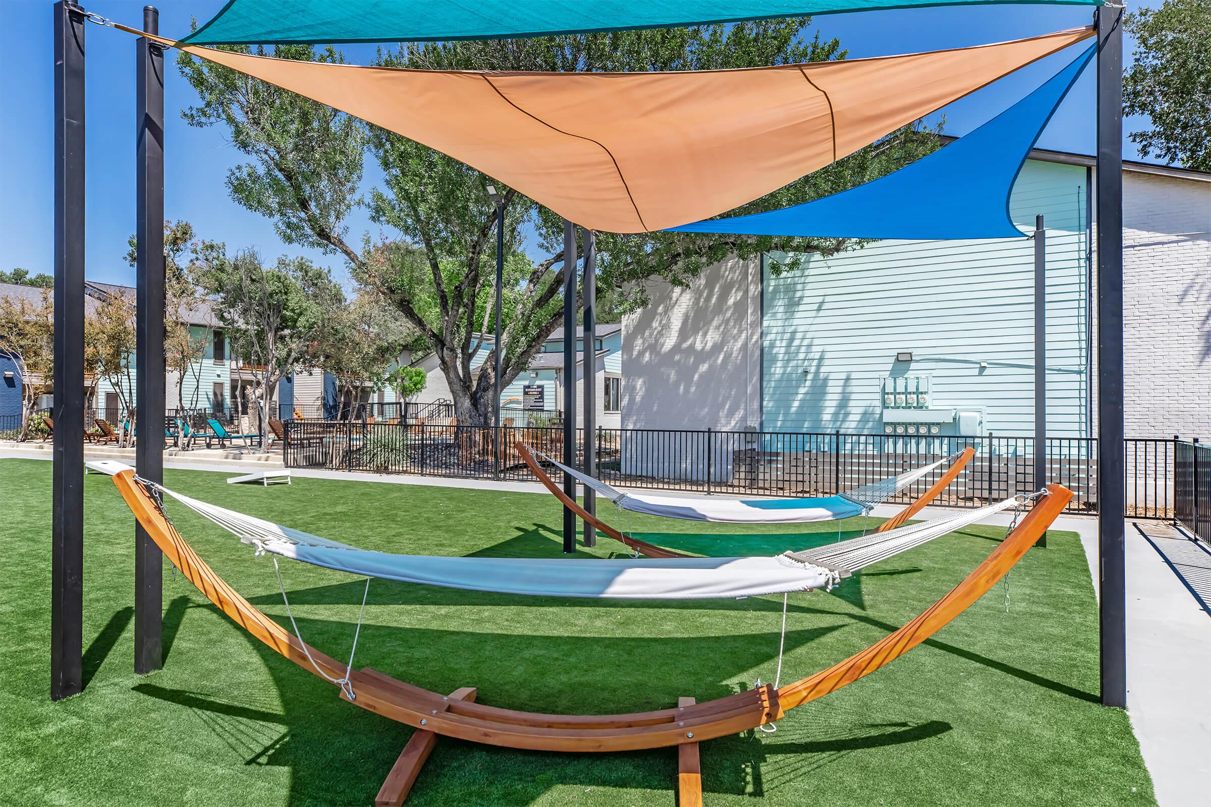 A relaxing outdoor space featuring three hammocks arranged under colorful shade sails. The hammocks are placed on artificial grass surrounded by trees and buildings, creating a peaceful environment for leisure and rest. Ideal for enjoying the outdoors in comfort.