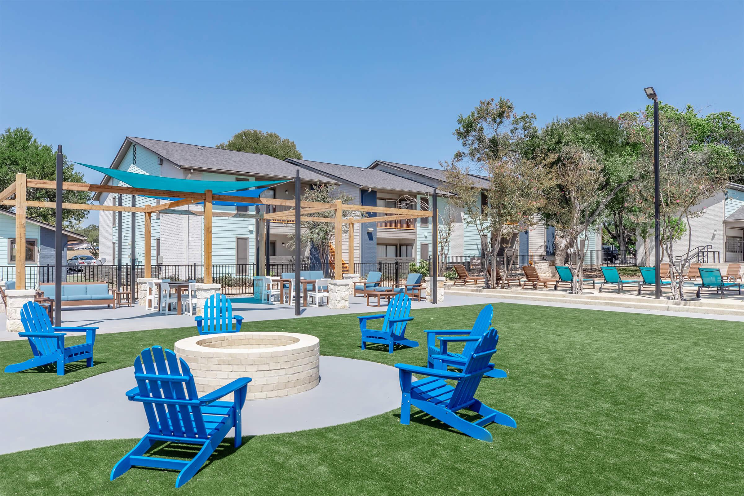 A sunny outdoor area featuring a circular fire pit surrounded by blue Adirondack chairs on green turf. In the background, there are modern buildings with balconies and a shaded lounging area with poolside chairs. Clear blue skies enhance the inviting atmosphere of the space.