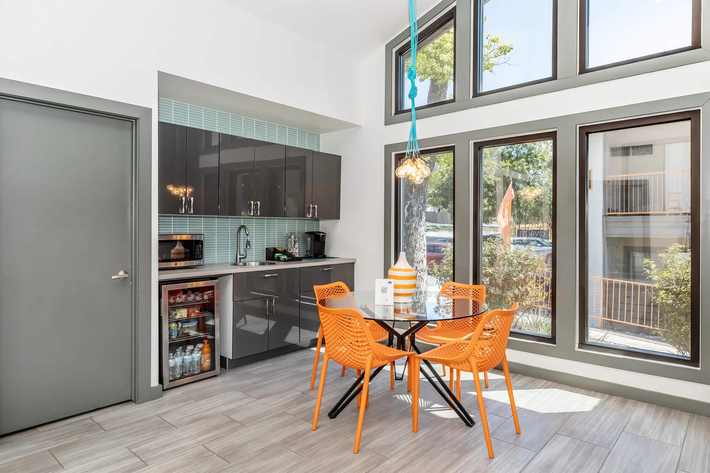 Modern kitchen space featuring a dark cabinetry, a small dining table with orange chairs, and large windows allowing natural light. The kitchen includes a mini fridge and a stylish light fixture, complemented by green tile accents. The overall design is clean and contemporary.