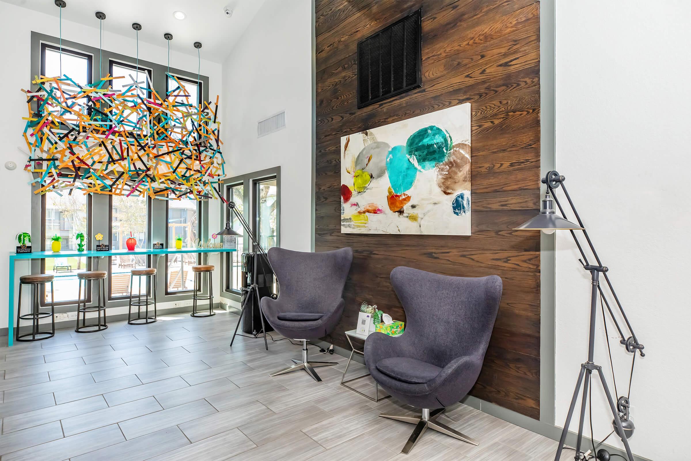 Modern interior space featuring two stylish gray chairs, a colorful abstract painting on a wooden accent wall, and a vibrant, artistic light fixture above. Large windows allow natural light, and a sleek bar area with colorful decorations is visible in the background.