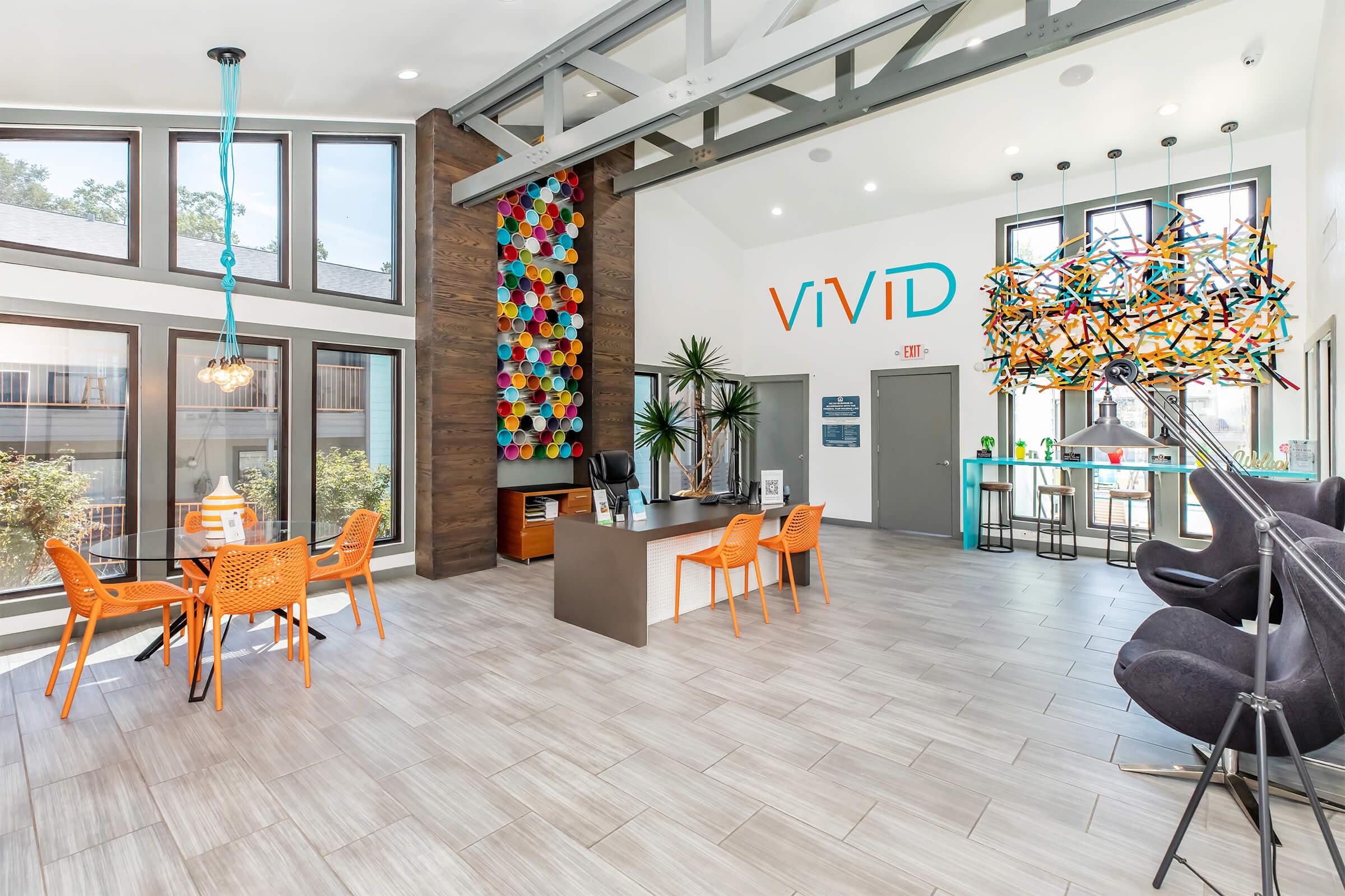 Bright, modern lobby with large windows and colorful artwork. Features include orange chairs around a table, a contemporary front desk, stylish lighting fixtures, and a vibrant wall installation. Lush green plants add a fresh touch to the inviting space.