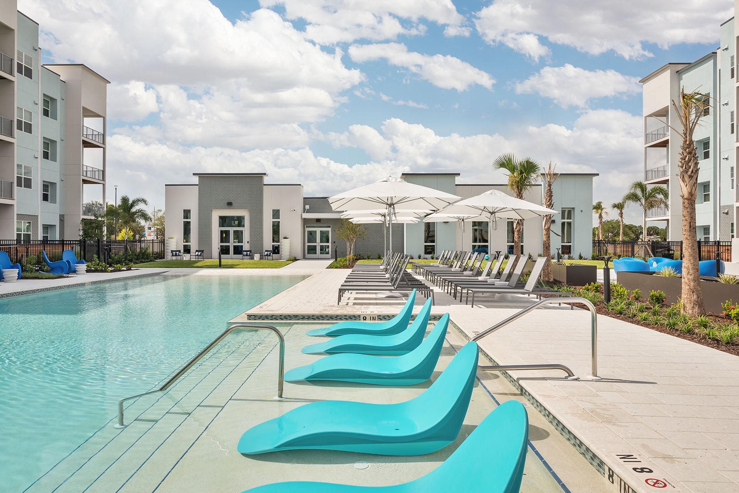 A modern outdoor pool area featuring turquoise lounge chairs, shaded umbrellas, and well-maintained landscaping. Surrounding the pool are contemporary apartment buildings under a partly cloudy sky, creating a relaxing atmosphere for residents and visitors.
