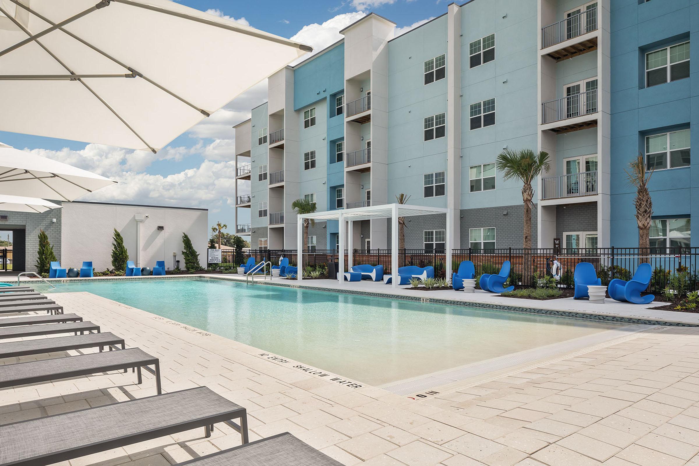 A modern outdoor swimming pool surrounded by lounge chairs and umbrellas, with blue residential buildings in the background. Palm trees are lined along the pool area, creating a tropical ambiance. The sky is clear with a few clouds, enhancing the relaxing atmosphere of the space.