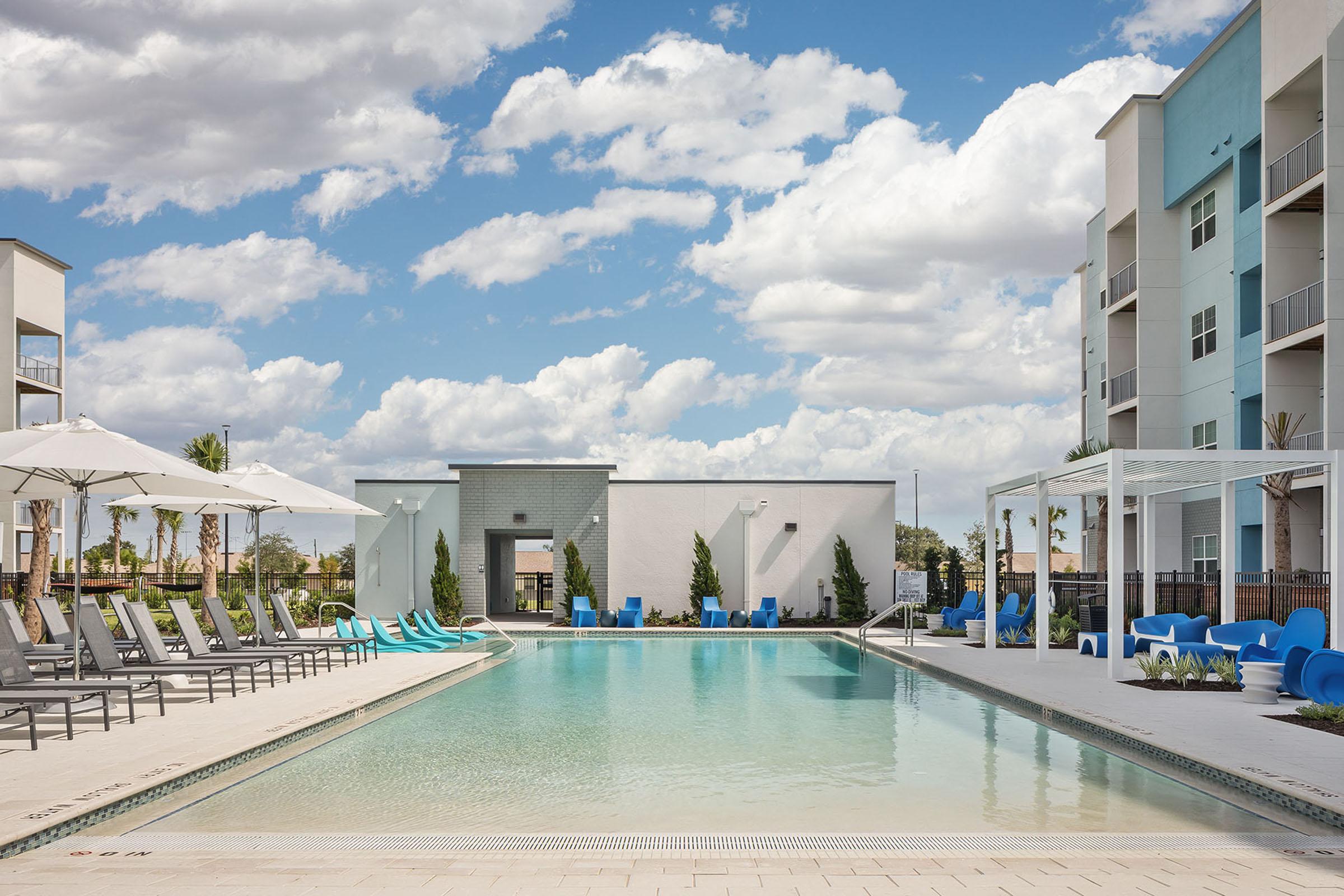 A modern swimming pool surrounded by lounge chairs and shaded areas, with tall buildings in the background. The sky is partly cloudy, adding to the inviting atmosphere of the outdoor space.
