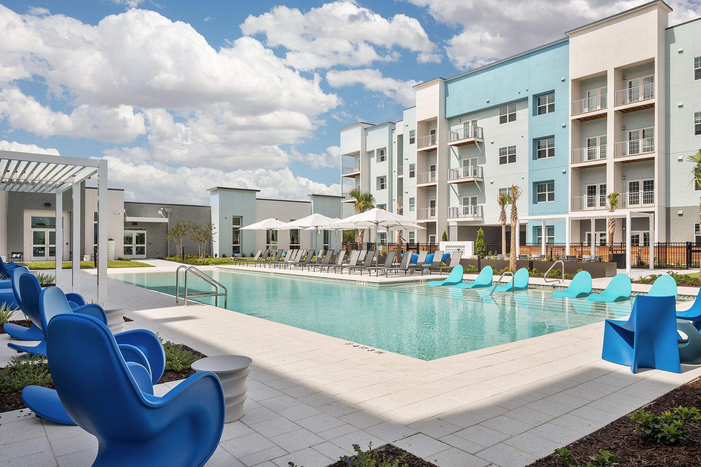 A modern outdoor swimming pool surrounded by lounge chairs and colorful blue seating. The pool area features umbrellas for shade and is set against a backdrop of contemporary apartment buildings. The sky is partly cloudy, creating a bright and inviting atmosphere.