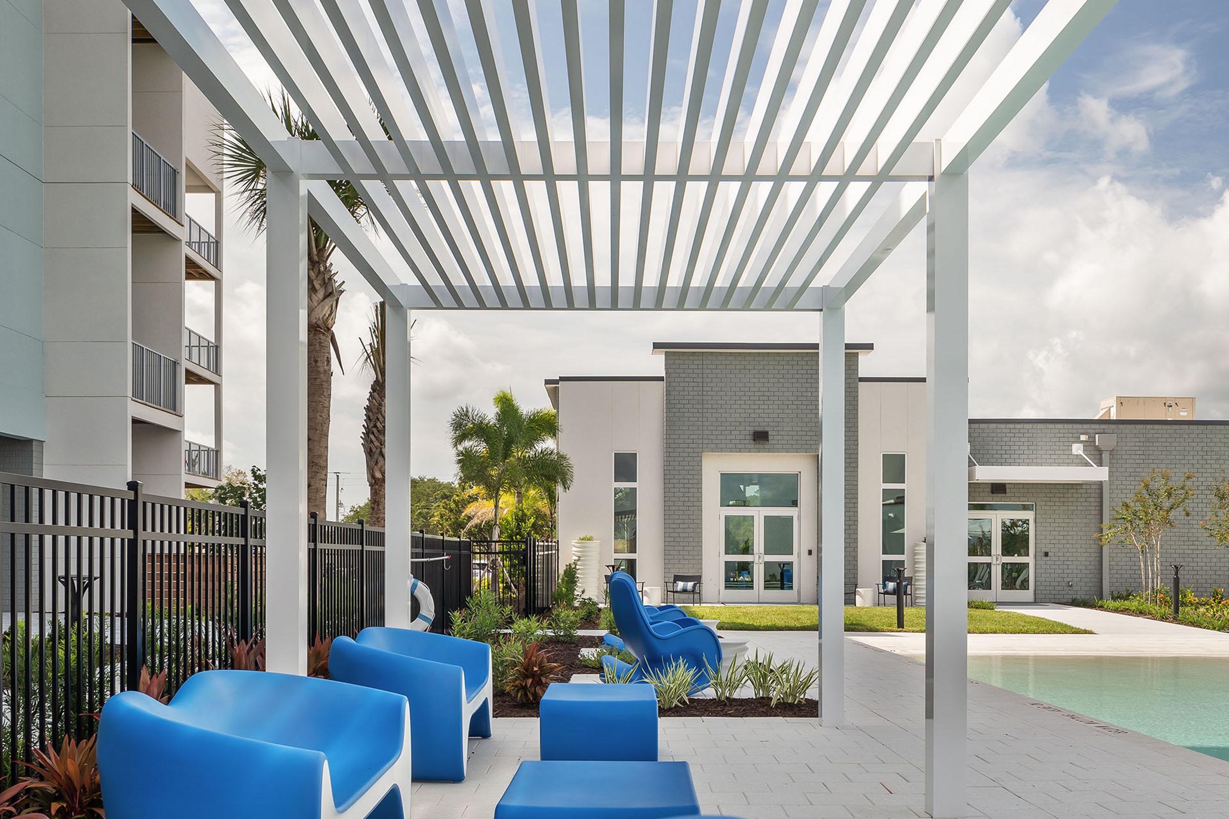 A modern outdoor area featuring a stylish pergola with horizontal slats, bright blue lounge chairs, and a clear swimming pool in the background. Lush greenery surrounds the pool area, while an apartment building with large windows and a community space can be seen in the distance.