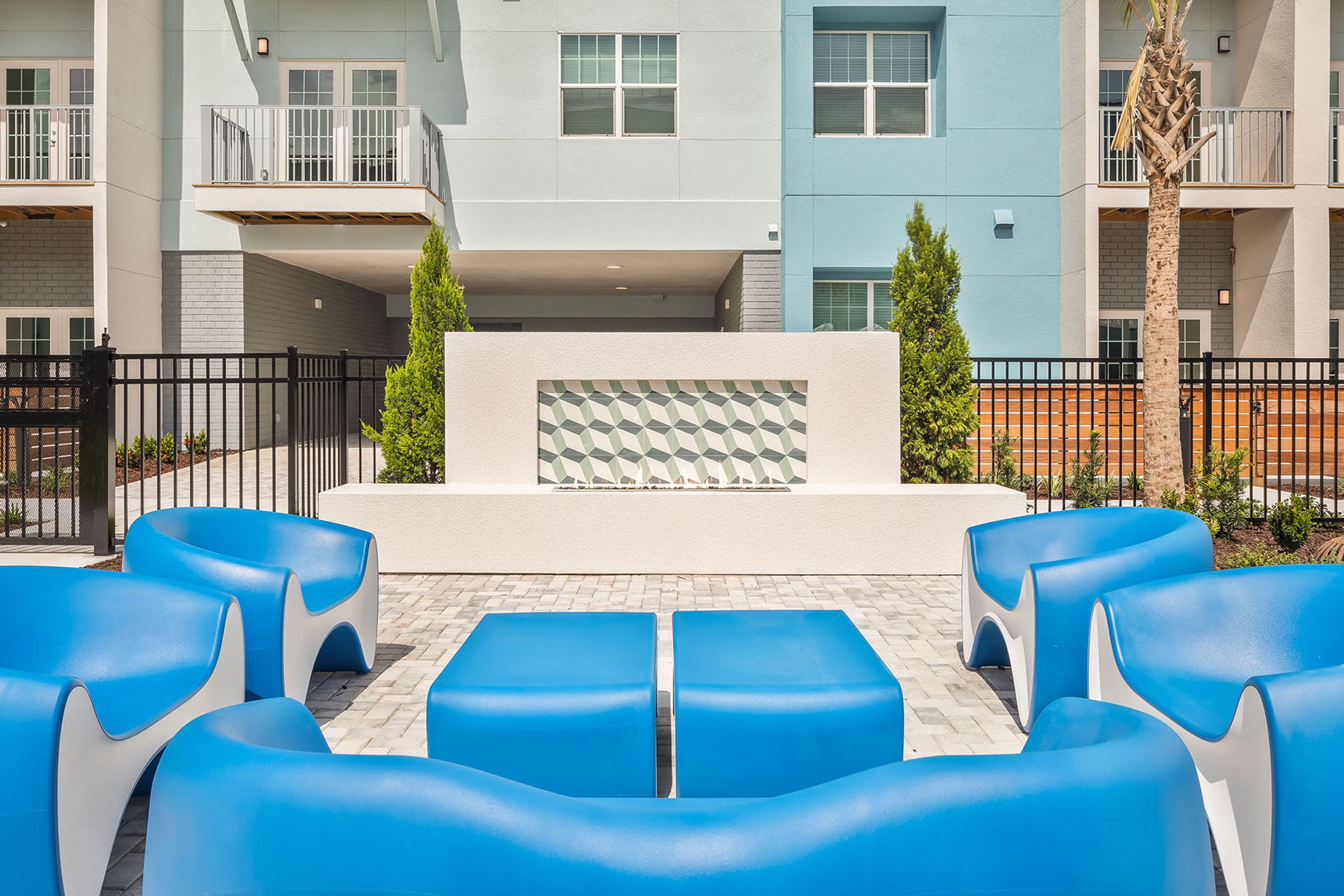 A modern outdoor seating area featuring blue chairs and tables arranged around a decorative water feature. The background includes multi-story building facades and neatly landscaped greenery, creating a relaxing and inviting space.