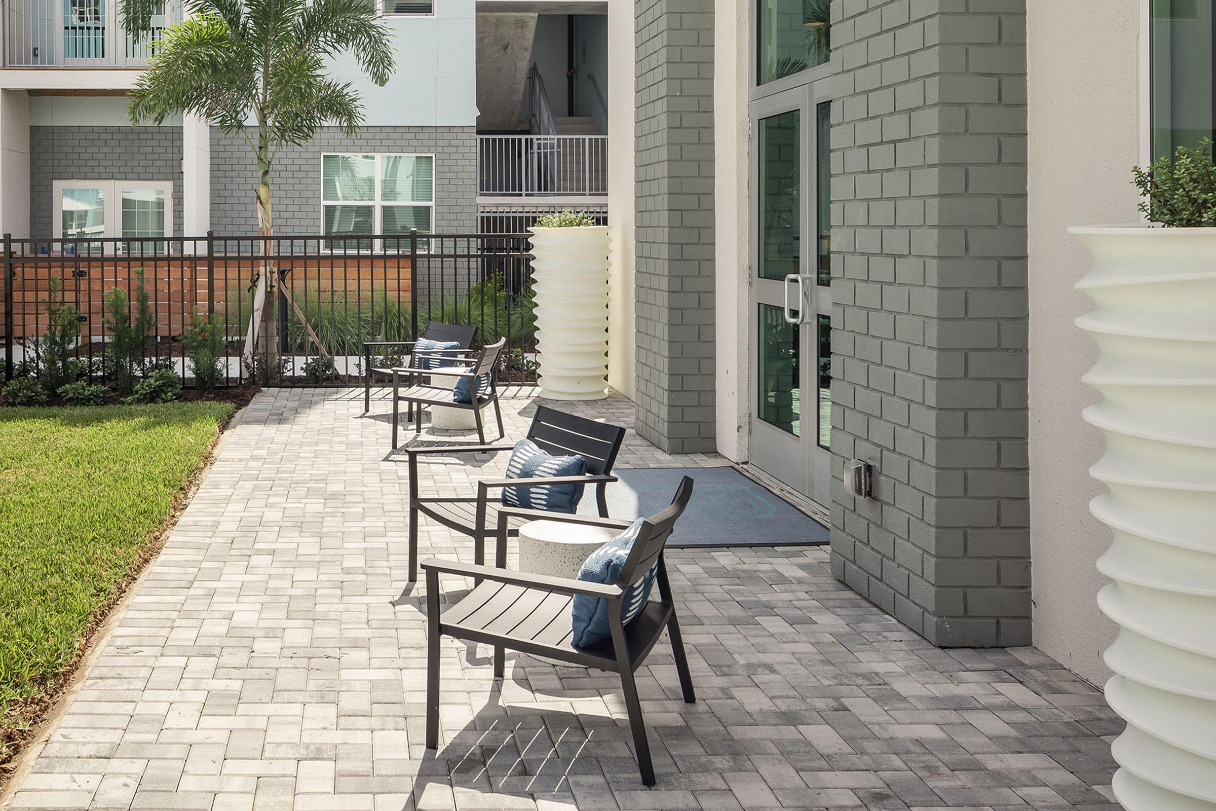 A modern outdoor patio area featuring several black lounge chairs with blue cushions arranged on a stone pavement. In the background, there are white and gray buildings with large windows, and a palm tree adds a tropical touch to the setting.