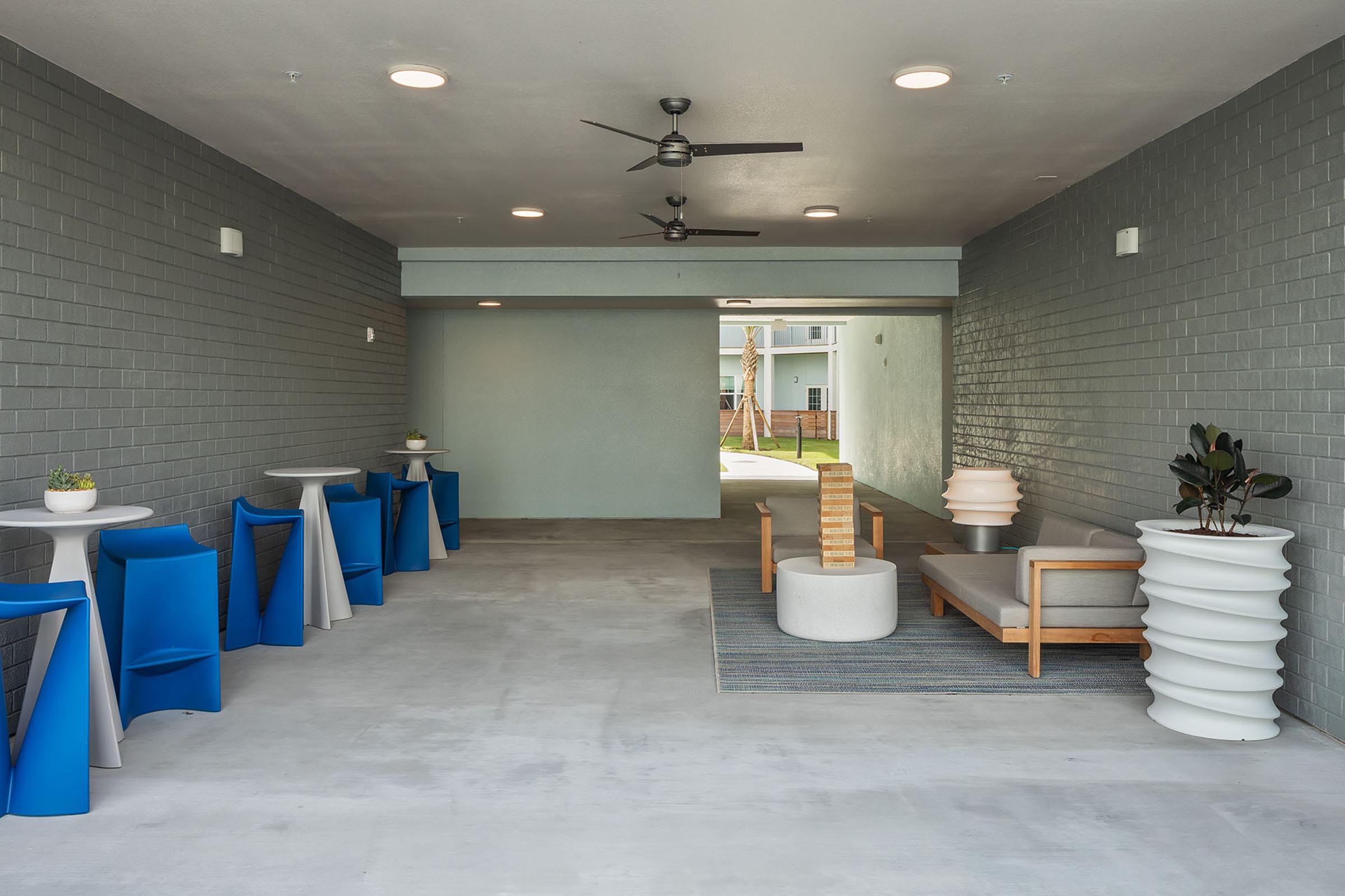 A modern, open lounge area featuring gray walls and concrete flooring. It includes seating with a gray sofa and a potted plant, along with white and blue tables and chairs. Ceiling fans are installed, and there is a view of an outdoor space through an archway at the far end.