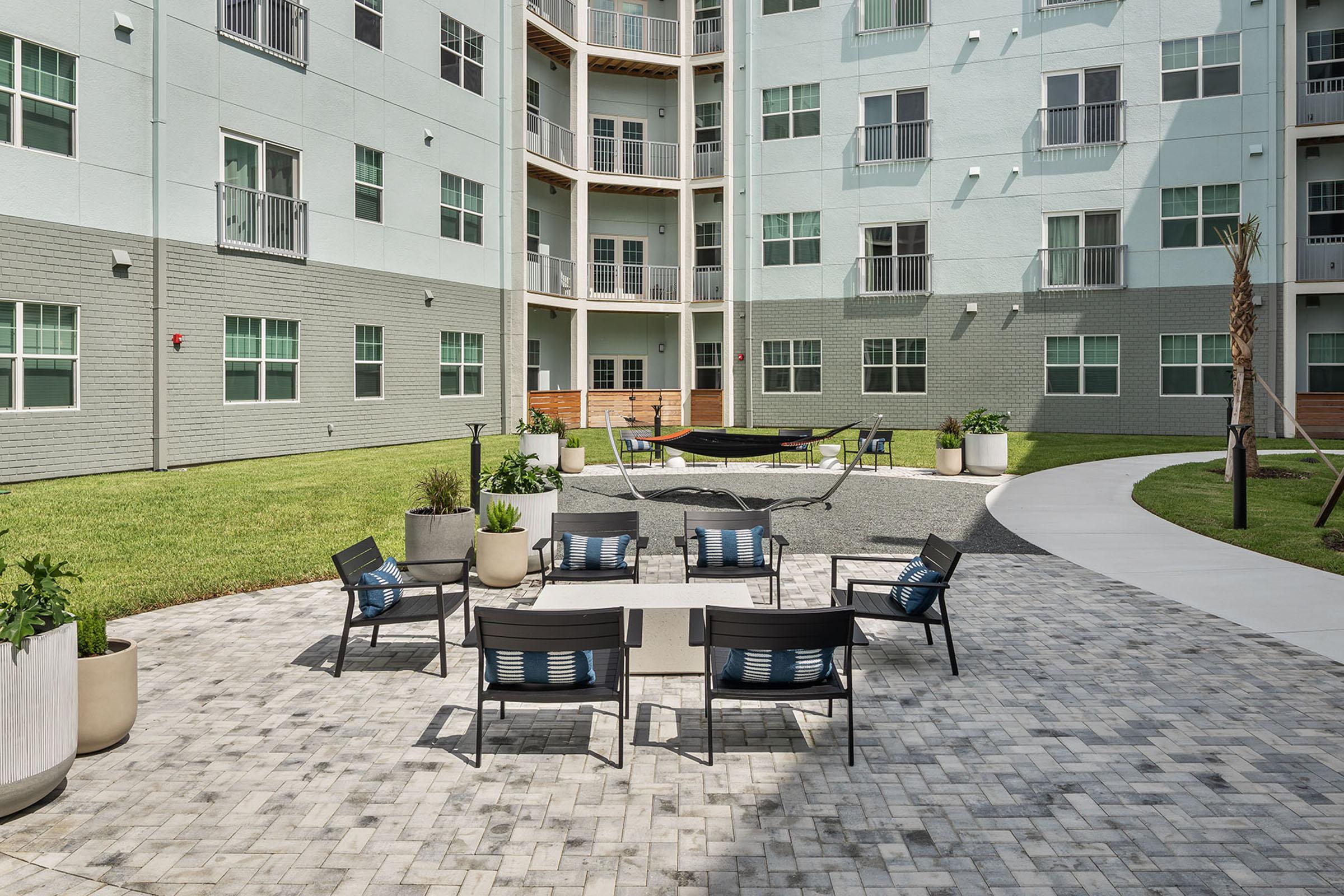 A modern outdoor courtyard with a stone-paved area featuring a table and several chairs. Lush green grass and potted plants surround the space. In the background, there's a multi-story building with large windows and balconies. A hammock is seen in the far corner, adding to the relaxing atmosphere.