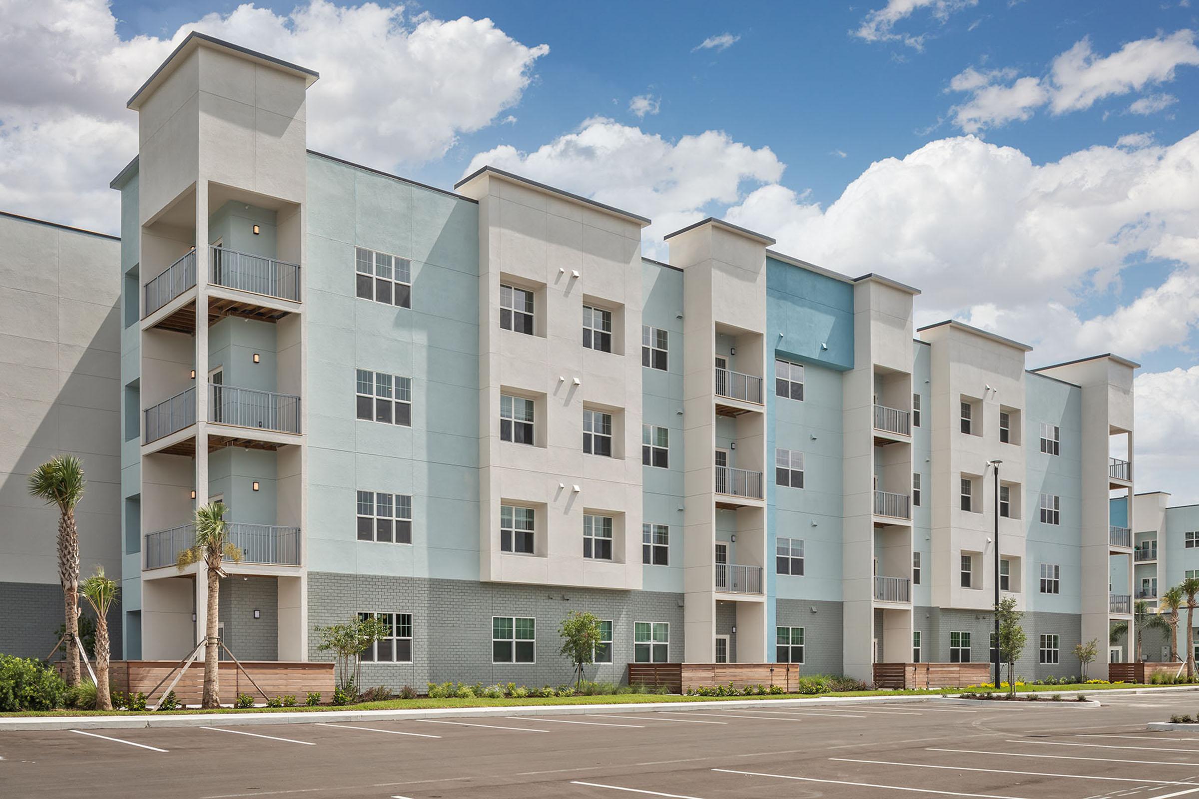 A modern apartment building with a light blue and white exterior, featuring multiple floors, balconies, and large windows. The surrounding area includes freshly landscaped greenery and parking spaces, set against a backdrop of a partly cloudy sky.