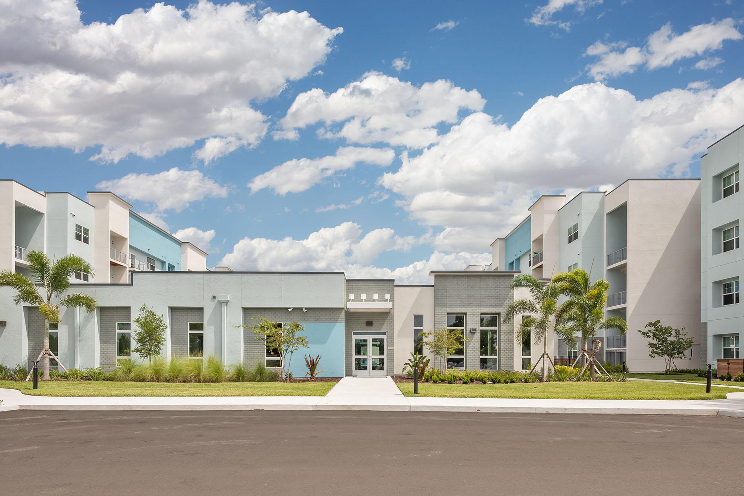 Modern residential building with a light blue and gray facade, surrounded by greenery. Clear blue sky with fluffy white clouds above. A landscaped area with palm trees is visible in the foreground, along with a paved walkway leading to the entrance of the building.