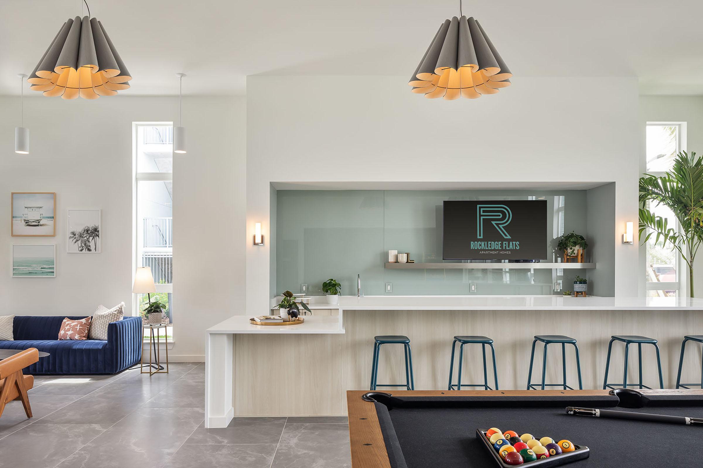 A modern lounge area featuring a stylish bar with high stools, a pool table, and contemporary light fixtures. There are decorative elements like plants and wall art, creating a welcoming atmosphere. A logo for "Rockledge Flats" is displayed on the back wall. Natural light fills the space through large windows.