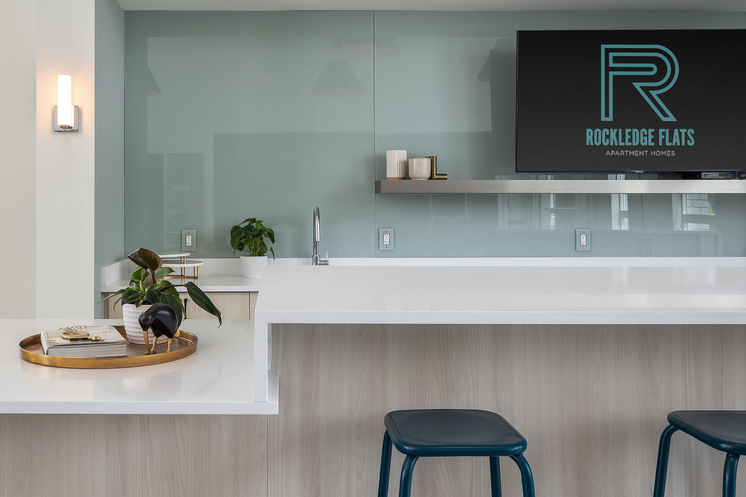 Modern kitchen area featuring a sleek white countertop, a decorative plant, and a stylish tray. Behind, there’s a TV displaying the "Rockledge Flats" logo. The space is bright and minimalist, with contemporary lighting and elegant decor accents.