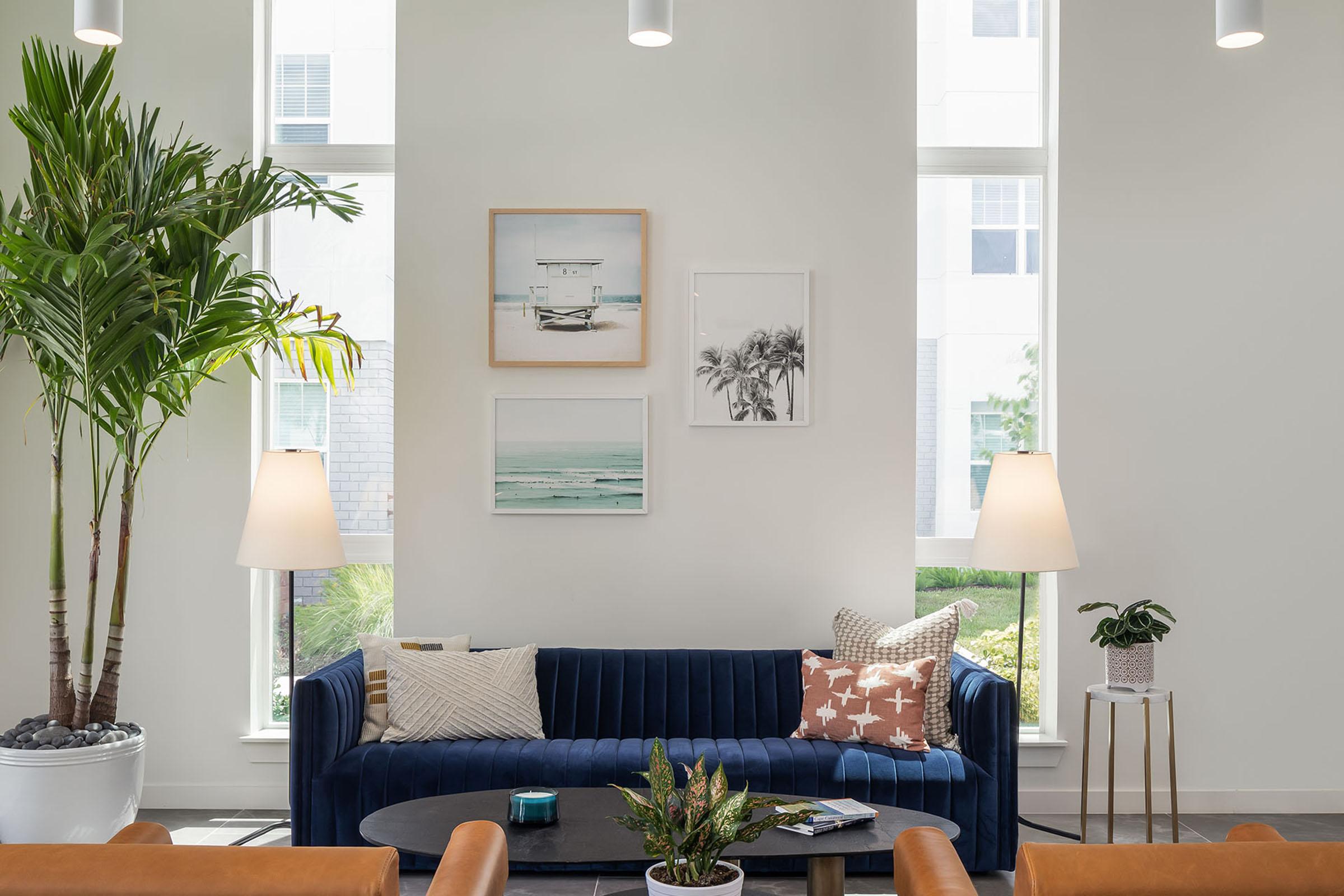 A modern, bright interior featuring a navy blue sofa adorned with patterned cushions, a round coffee table with plants and decor, two floor lamps, and potted greenery. The walls are decorated with framed beach-themed artwork, and large windows allow natural light to fill the space.