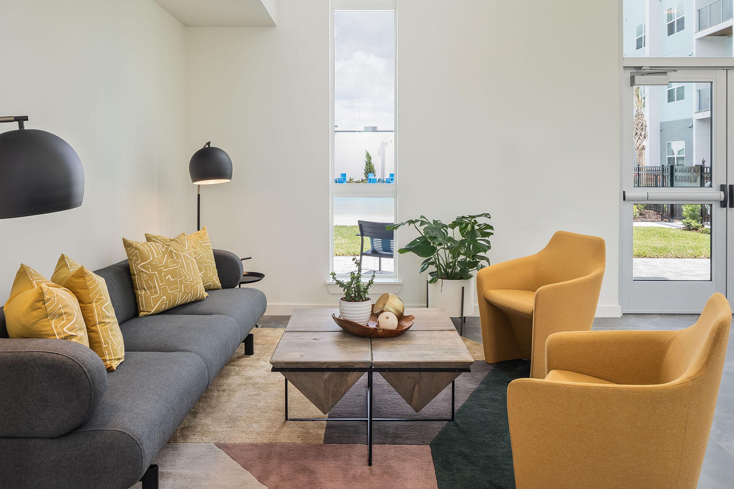 A modern living room featuring a gray sofa with yellow decorative pillows, a wooden coffee table with a plant and fruit bowl on it, and two vibrant yellow chairs. Large windows allow natural light to fill the space, with a view of an outdoor area visible. The decor includes a mix of textures and colors.